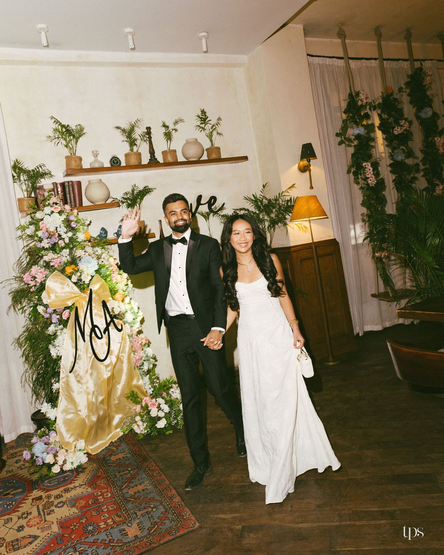 A couple in wedding attire holds hands and waves in a cozy room with floral arrangements, potted plants, warm lamps and a patterned rug.