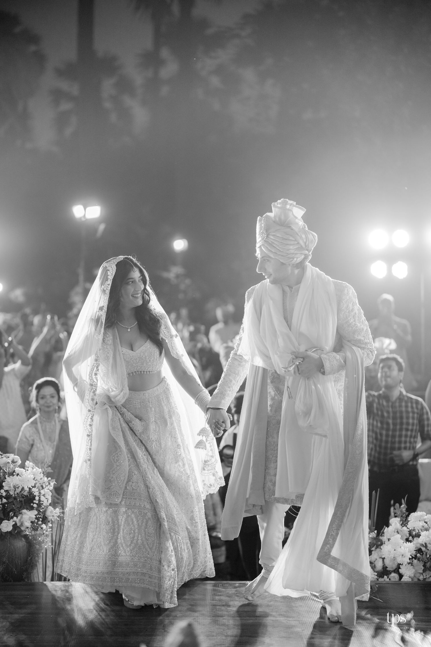 Black and white photo of Indian bride and groom in white attire walking together at an elegant wedding ceremony.