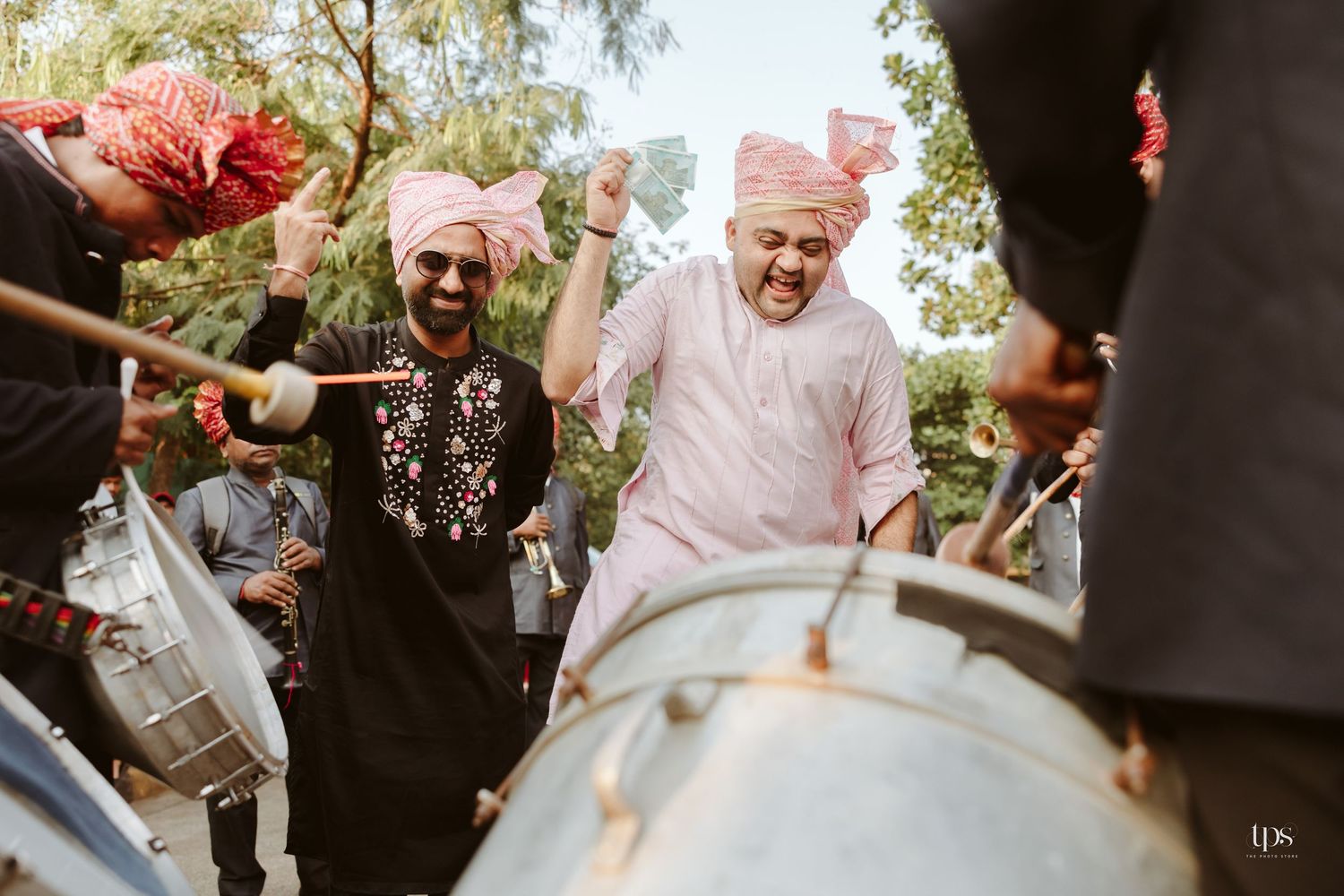 Two men in pink turbans celebrate joyfully with dhol drummers during a vibrant Indian wedding baraat ceremony.