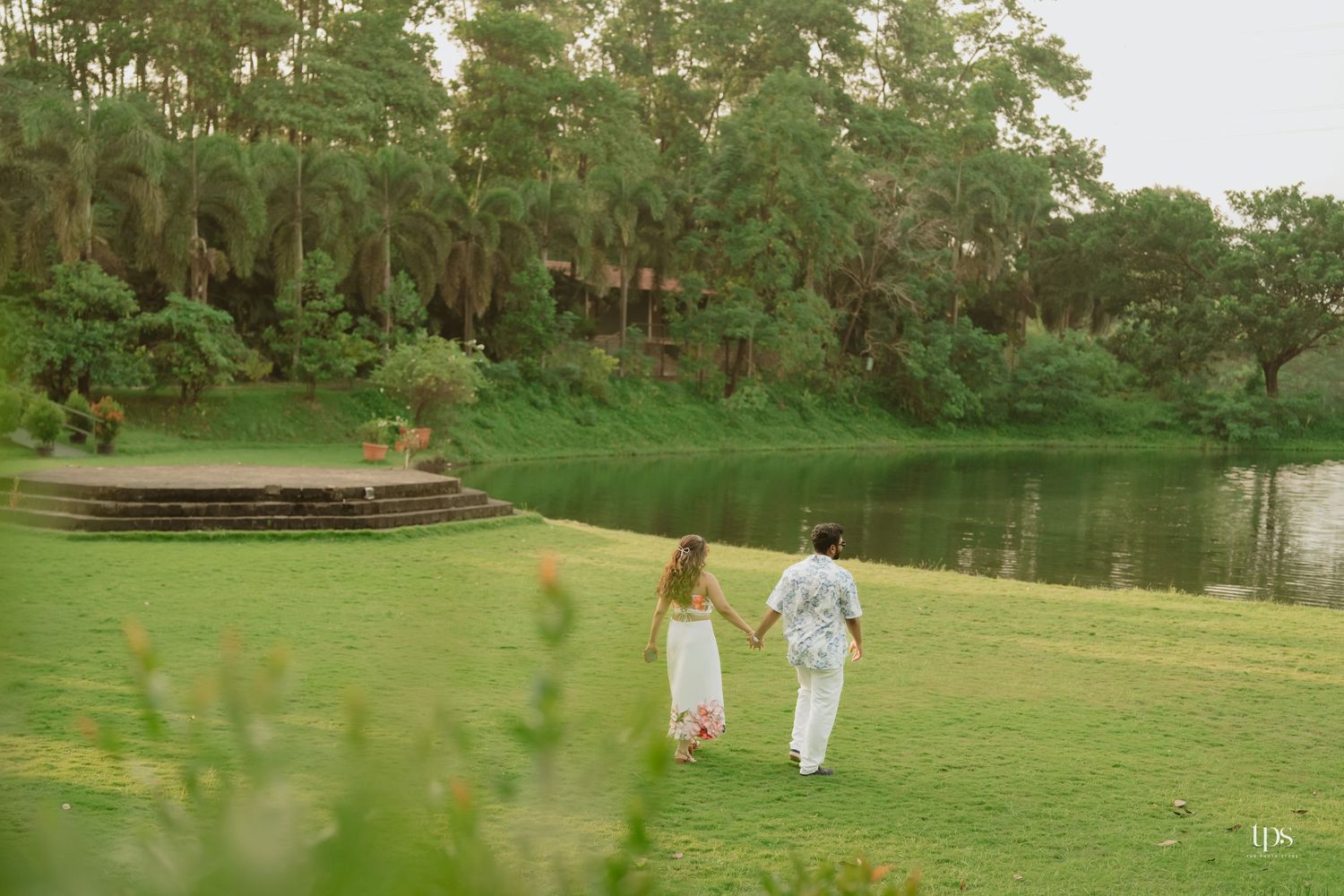 Two figures in white walking hand in hand near a serene lake surrounded by lush greenery.