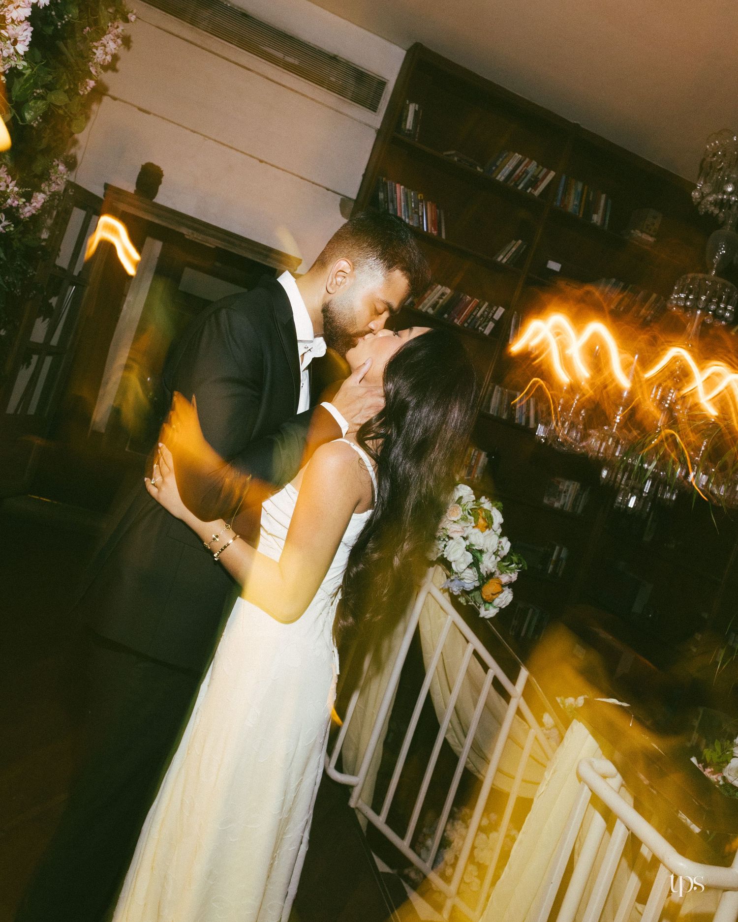 A couple in formal wedding attire share a kiss in a warmly lit room with bookshelves, white flowers, and glowing golden light streaks.