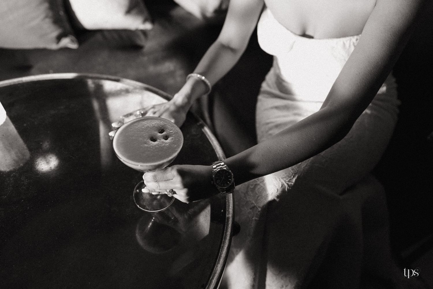 Black and white photo of a bride in a strapless gown pouring champagne into a glass on a round table.