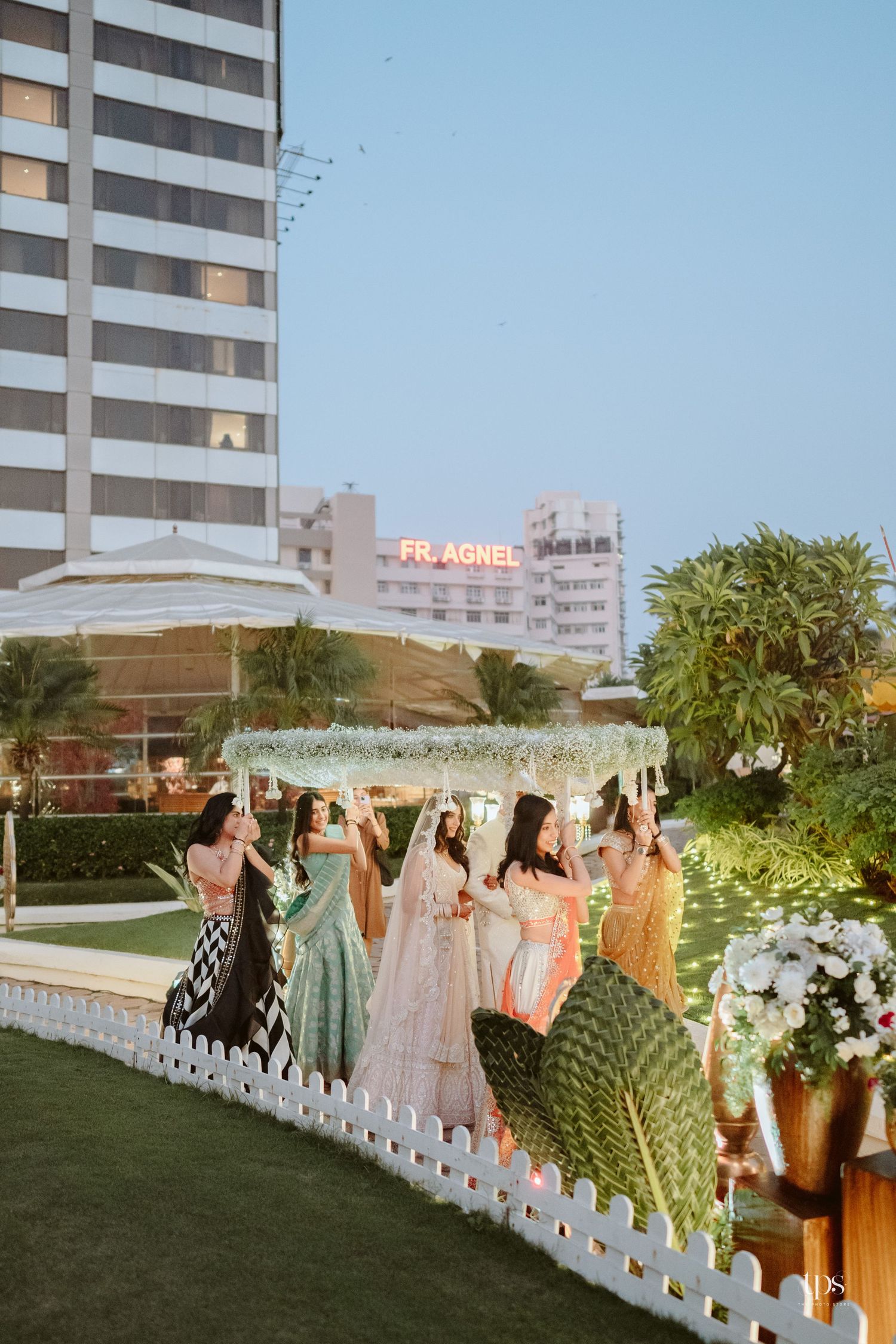 Bridesmaids in colorful lehengas stand under a floral mandap at an outdoor Indian wedding venue at dusk.