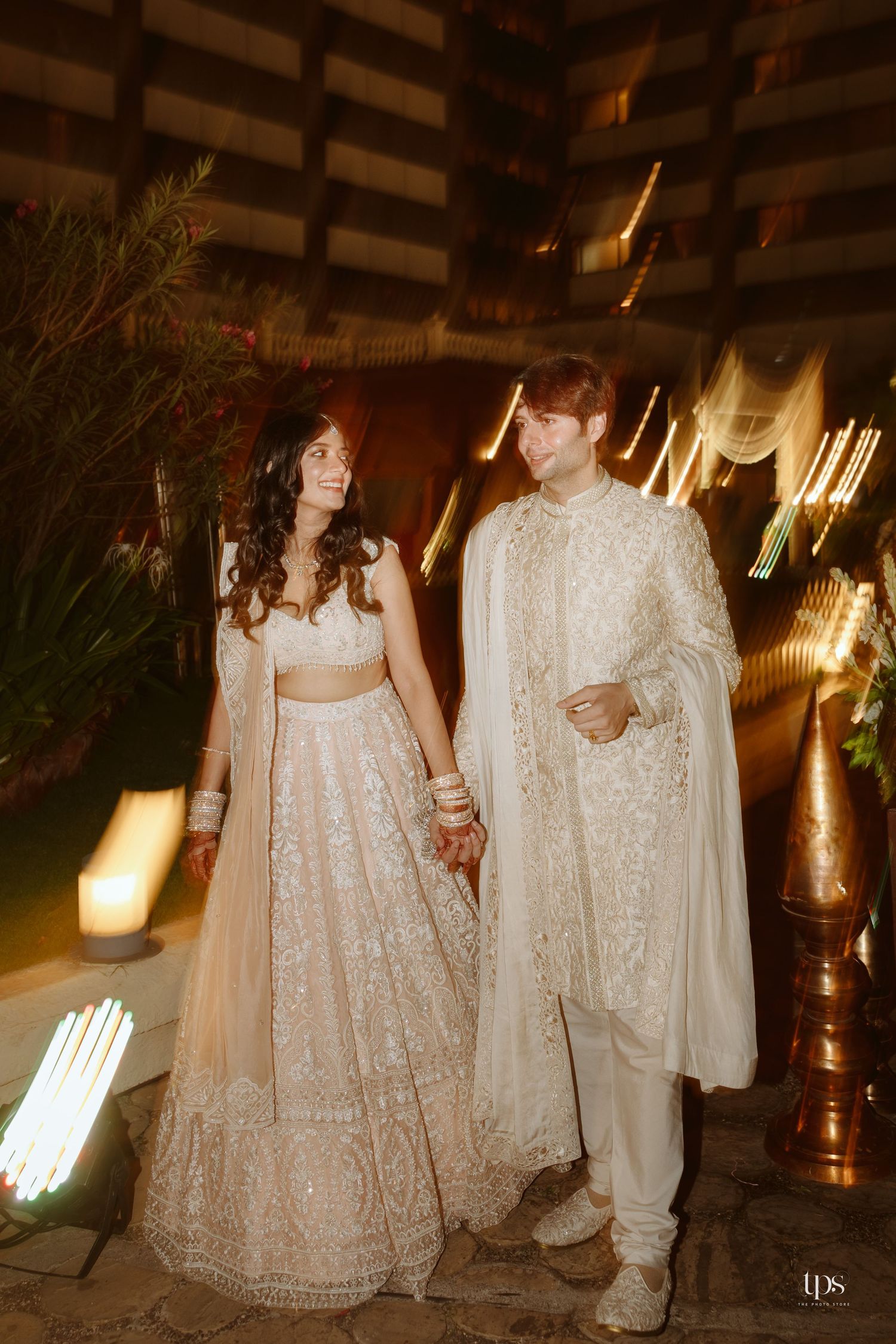 Couple in white Indian wedding attire, bride in embroidered lehenga, groom in sherwani with dupatta, posing at evening venue.
