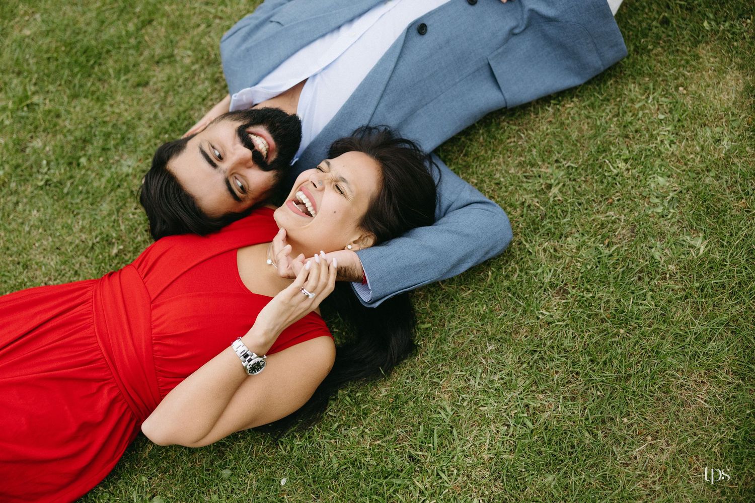 Couple lying on green grass, woman in red dress and man in blue suit, sharing a romantic outdoor moment.