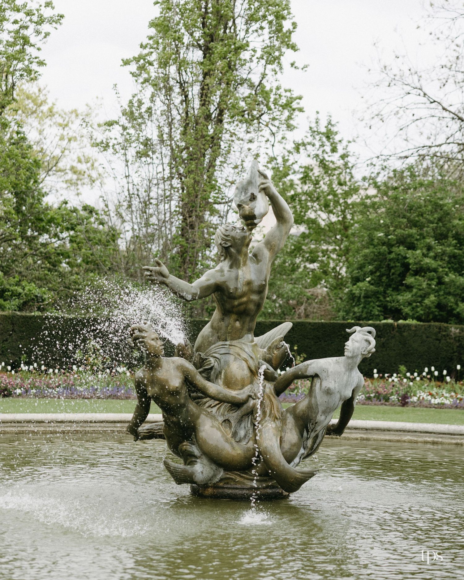 Classical stone fountain sculpture with figures and horses, surrounded by lush green trees in a formal garden setting.