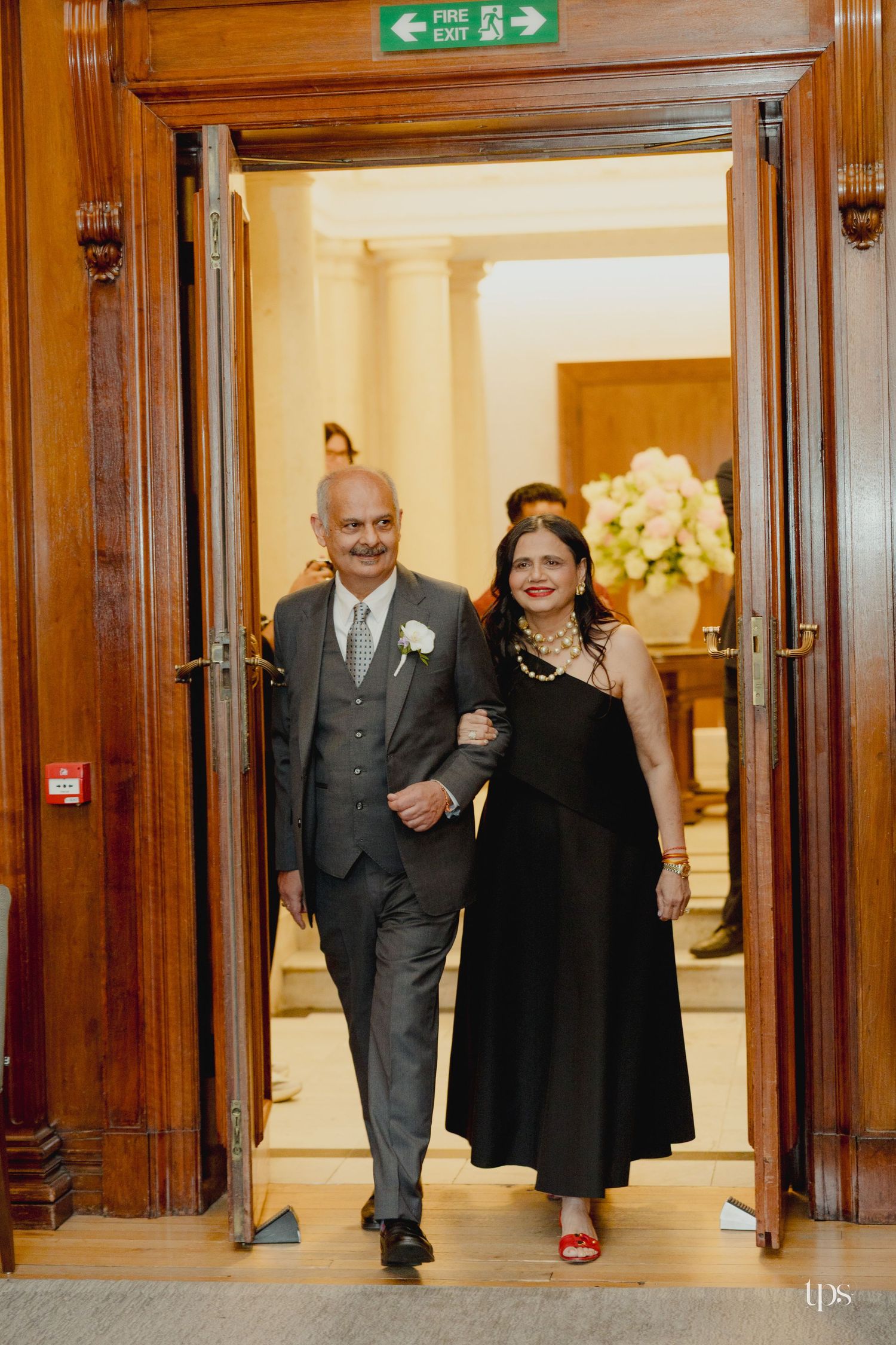 Elegant couple walking through ornate wooden doors at a formal event, man in gray suit, woman in black dress.