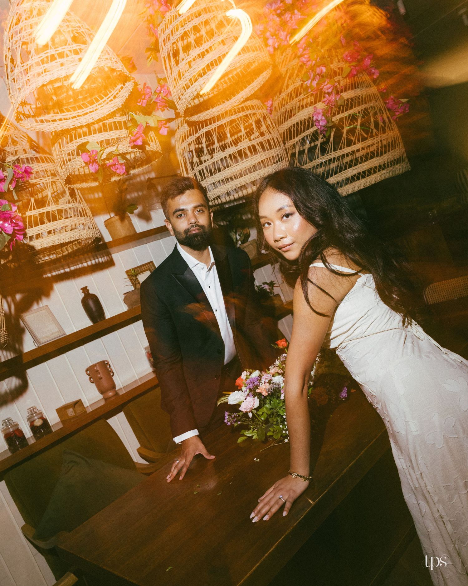 Bearded groom in a dark suit and bride in a white gown with a bouquet pose at a wooden table beneath hanging wicker lanterns.