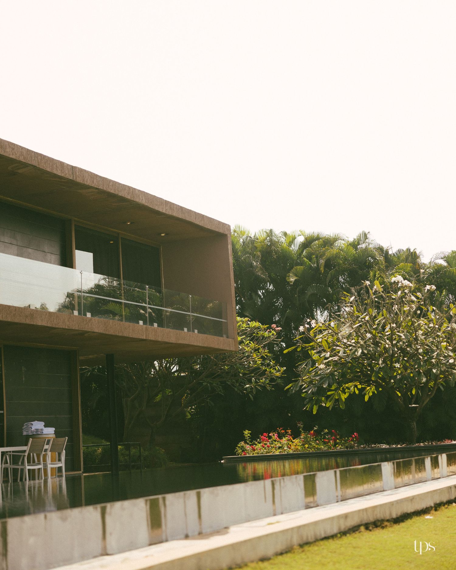 Modern two-story concrete house with glass balcony railings beside a reflecting pool, lush green lawn, and tropical trees under a hazy sky.