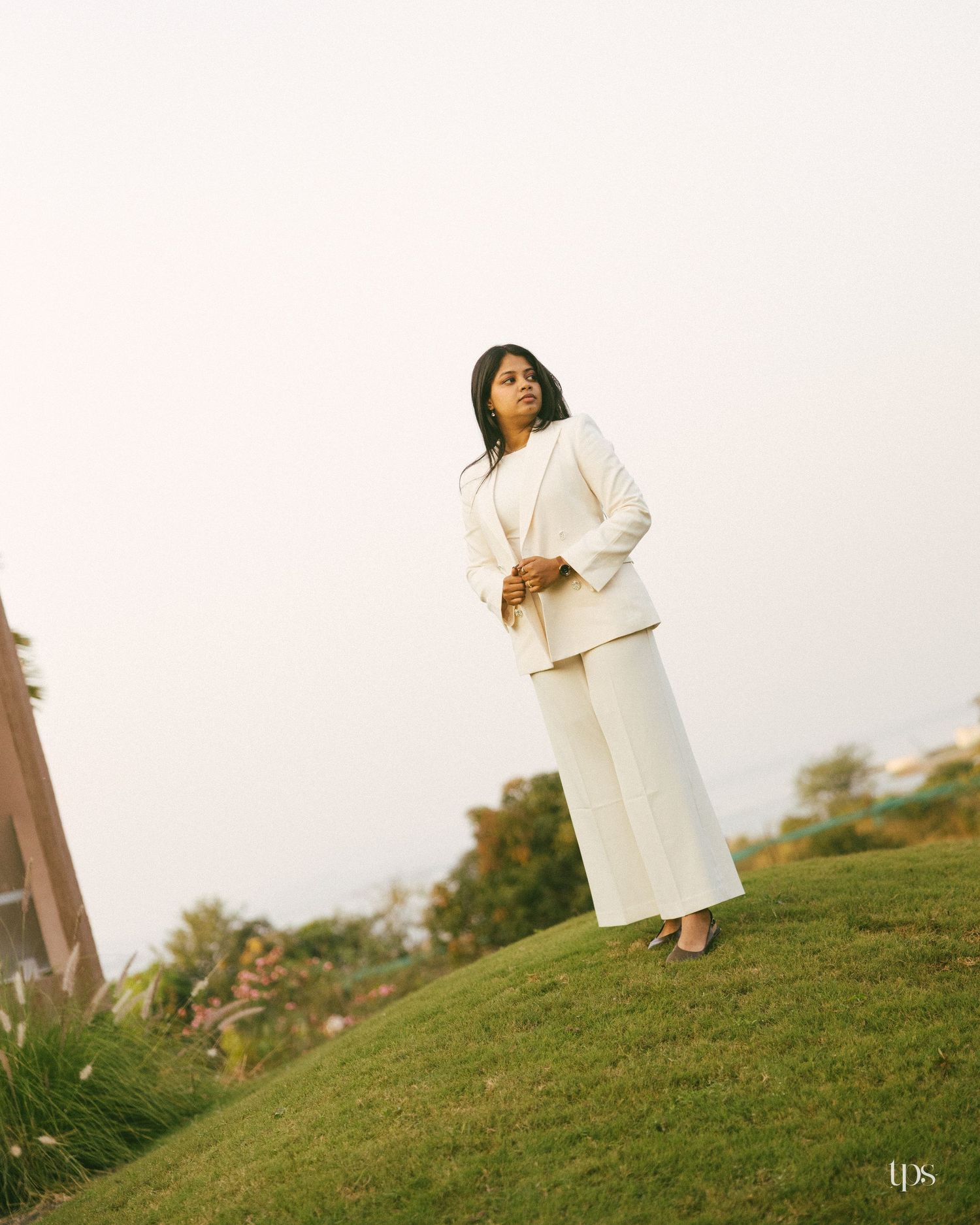 Woman in a white blazer and wide-leg trousers stands on a grassy hill, gazing into the distance under a soft, hazy sky.
