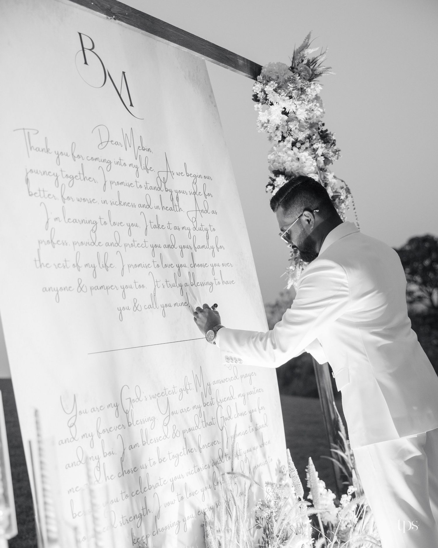 Groom in a white suit writes on a large handwritten vows sign beside a floral arch at an outdoor wedding, in black and white.