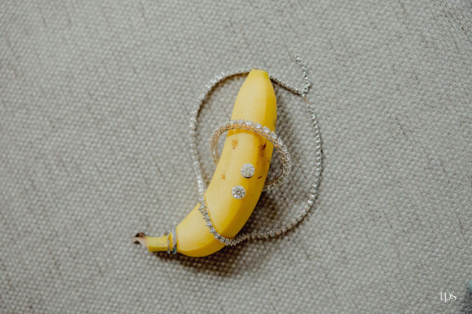 A yellow banana wrapped in a delicate diamond chain necklace, placed on a neutral textured surface.