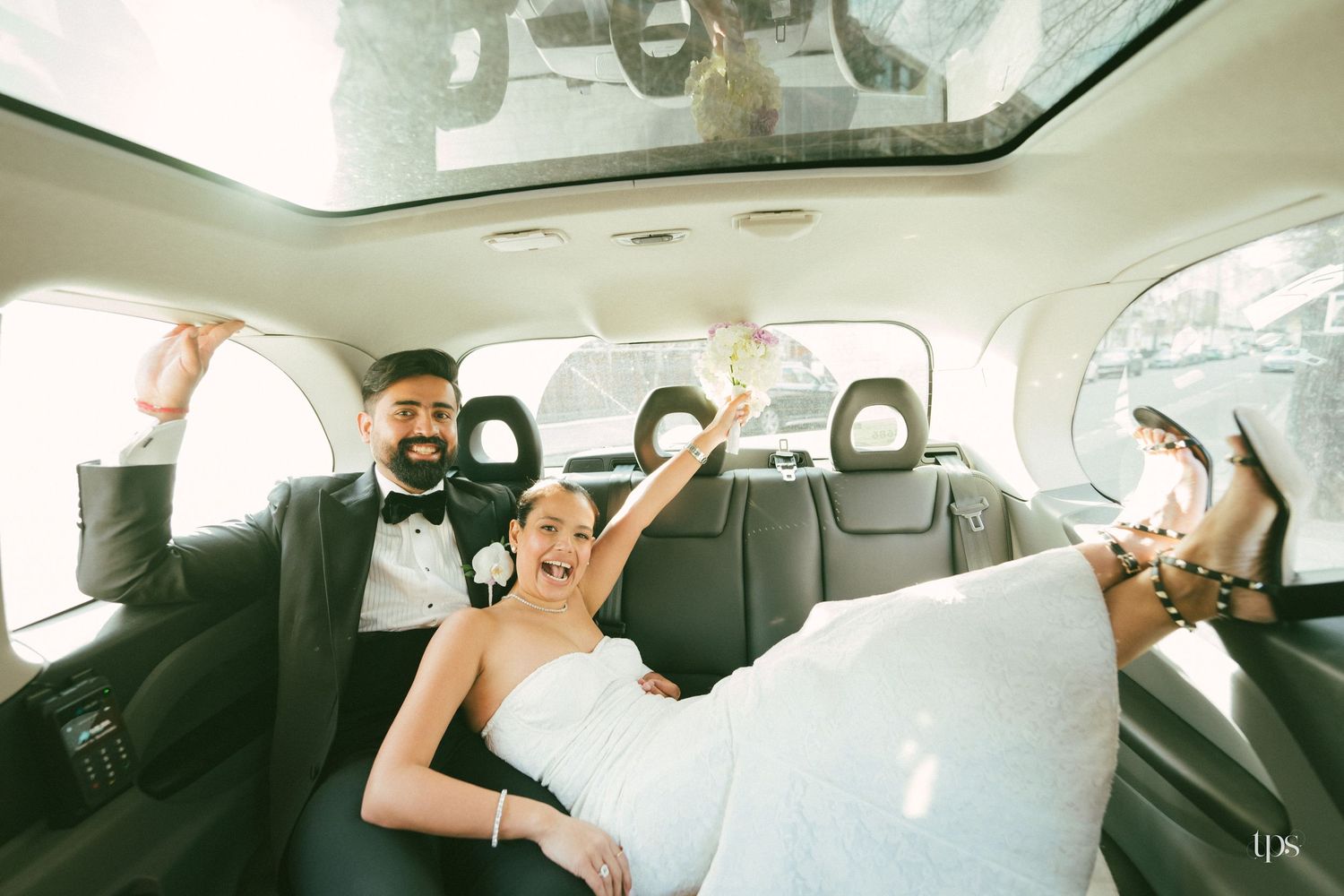 Joyful newlyweds celebrating in luxury car backseat, bride in white gown raising bouquet, groom in tuxedo smiling.
