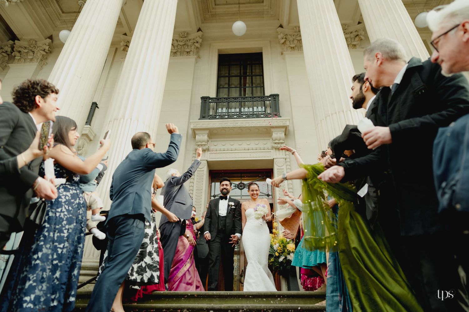 Bride and groom celebrate on grand building steps surrounded by cheering wedding guests in elegant attire.