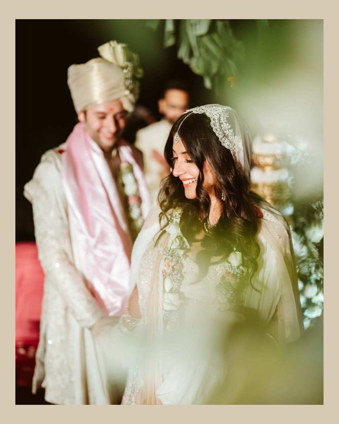 Bride in white embroidered lehenga with veil smiles at Indian wedding ceremony, groom in cream sherwani behind her.