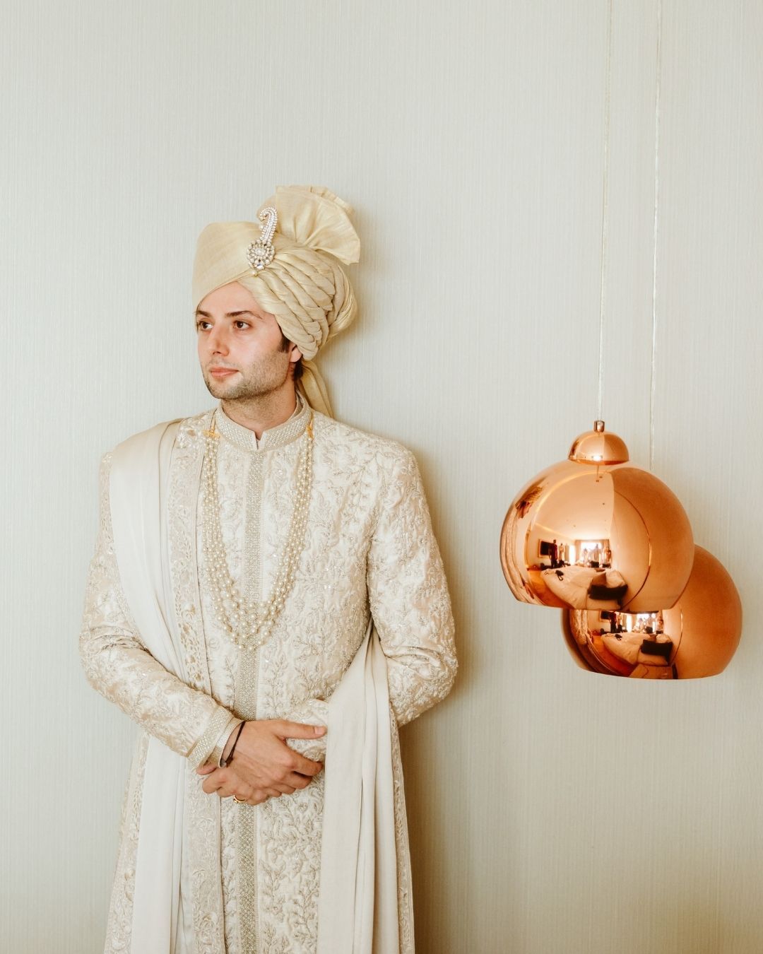 Groom in ivory embroidered sherwani and cream turban stands beside copper pendant lights.