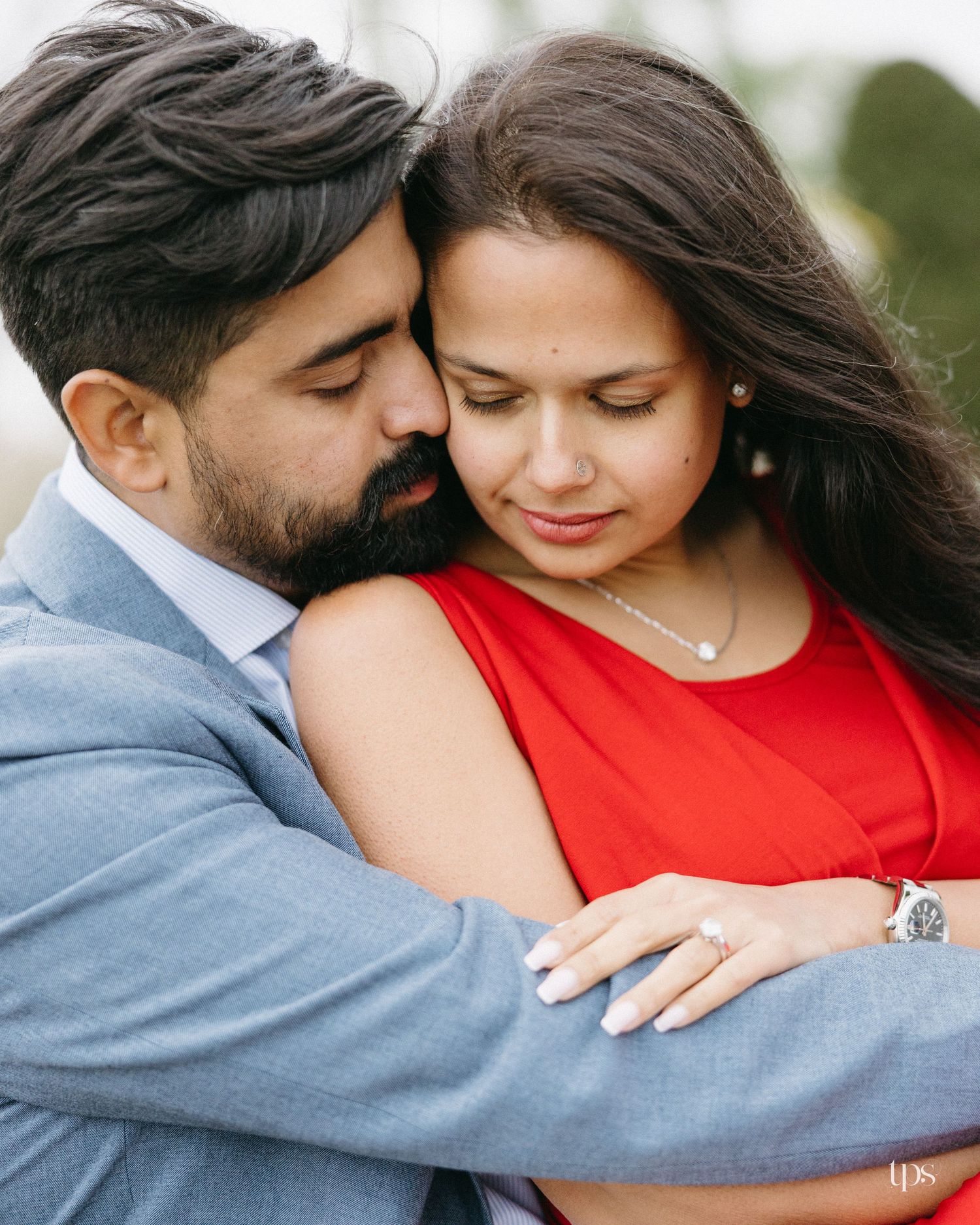 A couple in an intimate embrace, man in blue suit and woman in red dress, sharing a tender romantic moment outdoors.