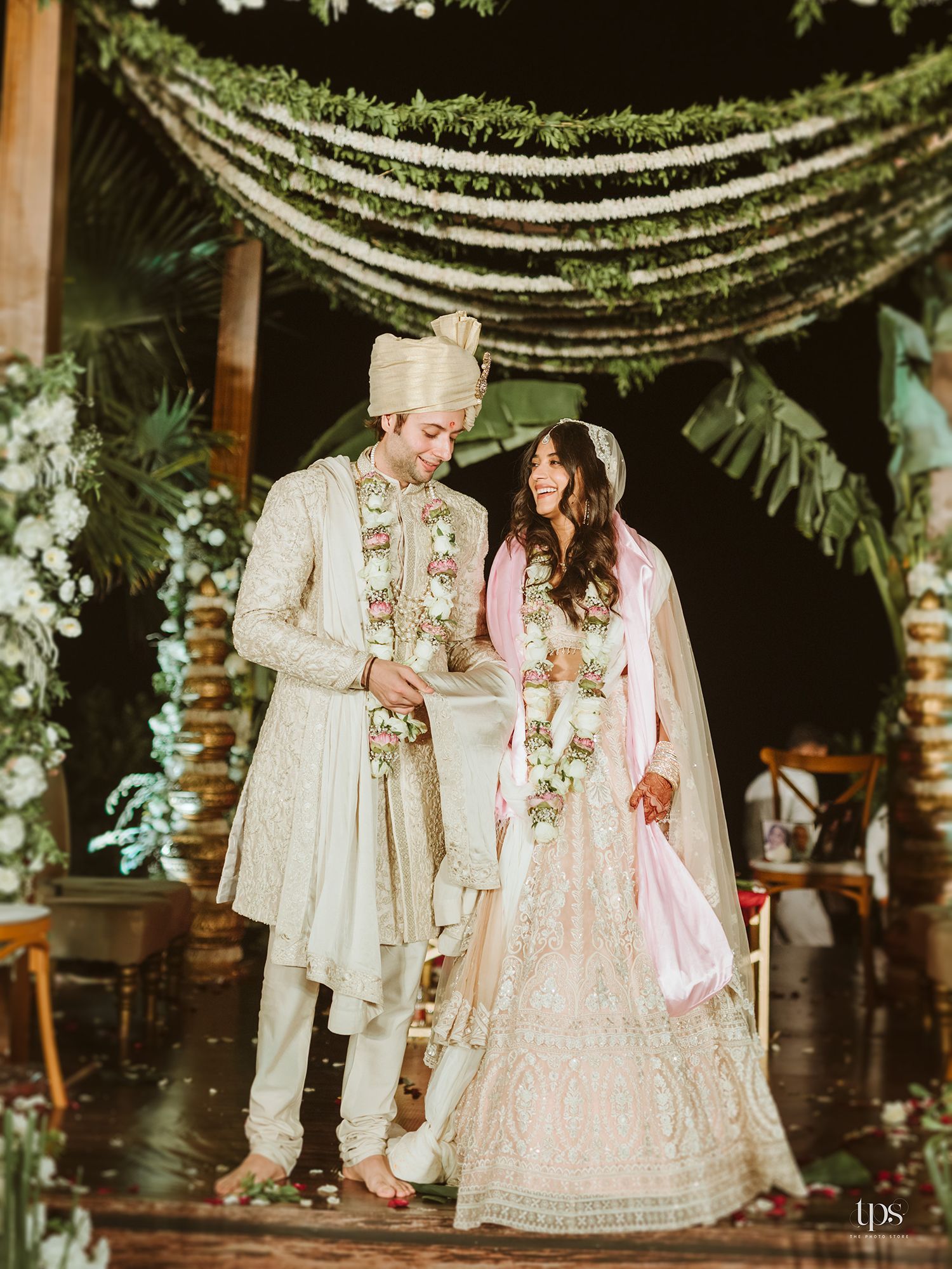 Couple in traditional Indian wedding attire stand under floral arch, bride in pink lehenga and groom in cream sherwani.