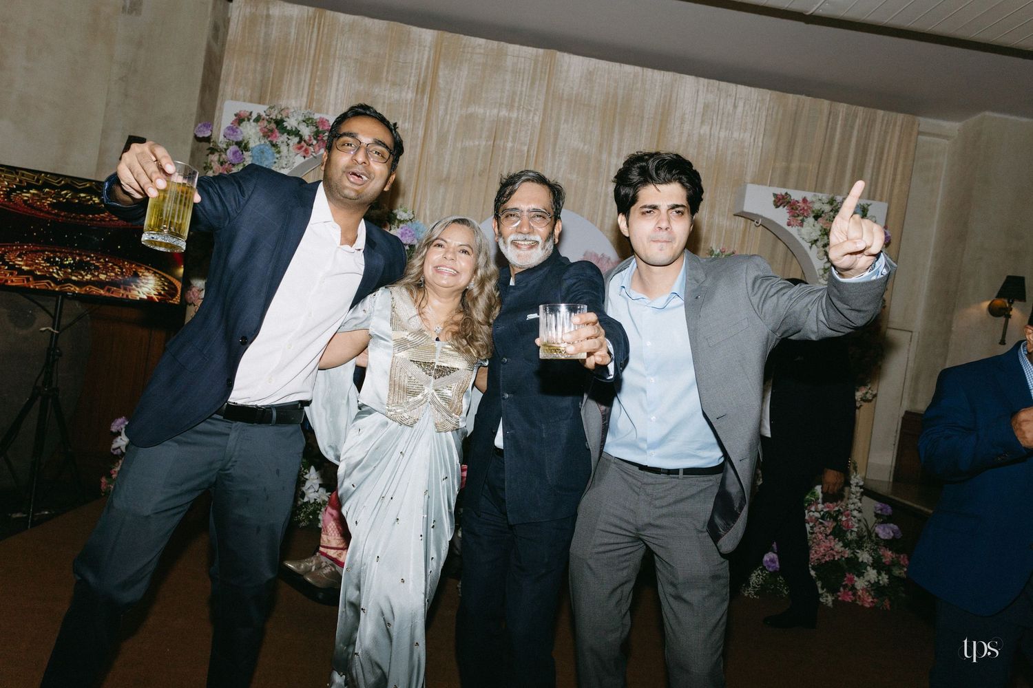 Four smiling guests pose together at a festive indoor party, holding drinks, with one man raising his hand in a cheerful gesture.