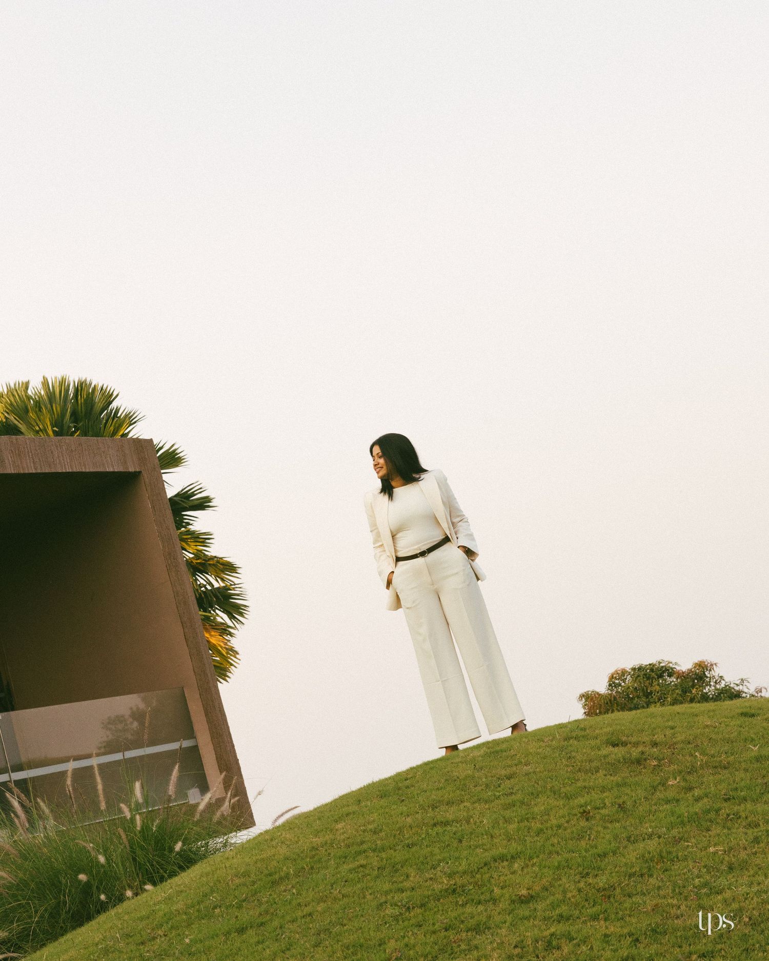 Woman in a white blazer and wide-leg trousers stands on a grassy hill beside a modern brown structure and palm tree under a soft hazy sky.