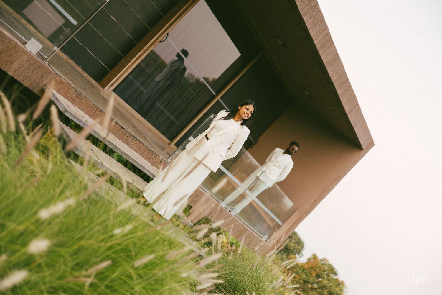 Two people in white outfits stand on a glass-railed balcony of a modern brick building, with tall grass in the foreground.