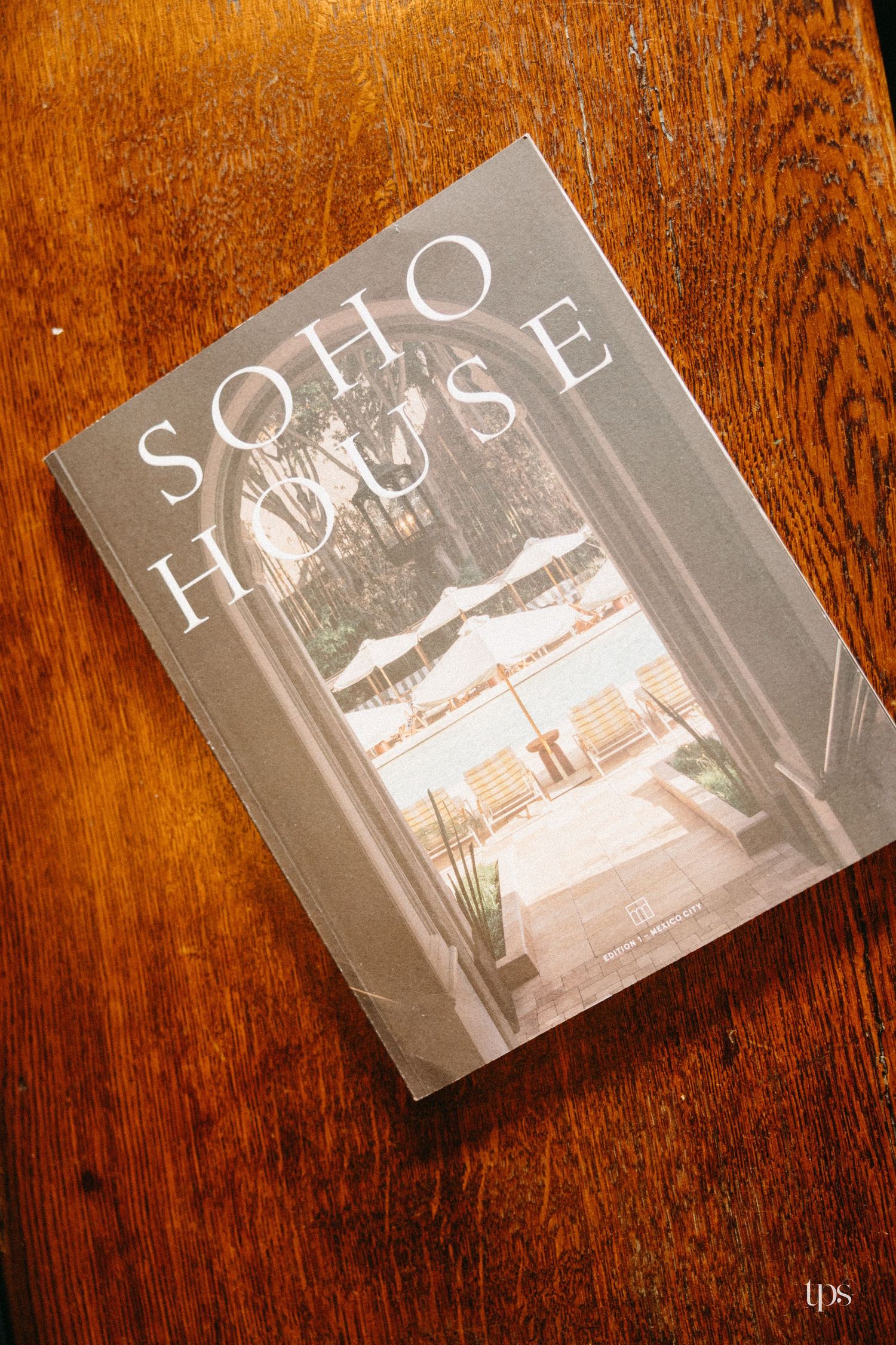 Soho House book resting on a wooden table, featuring a serene poolside scene on its gray cover.