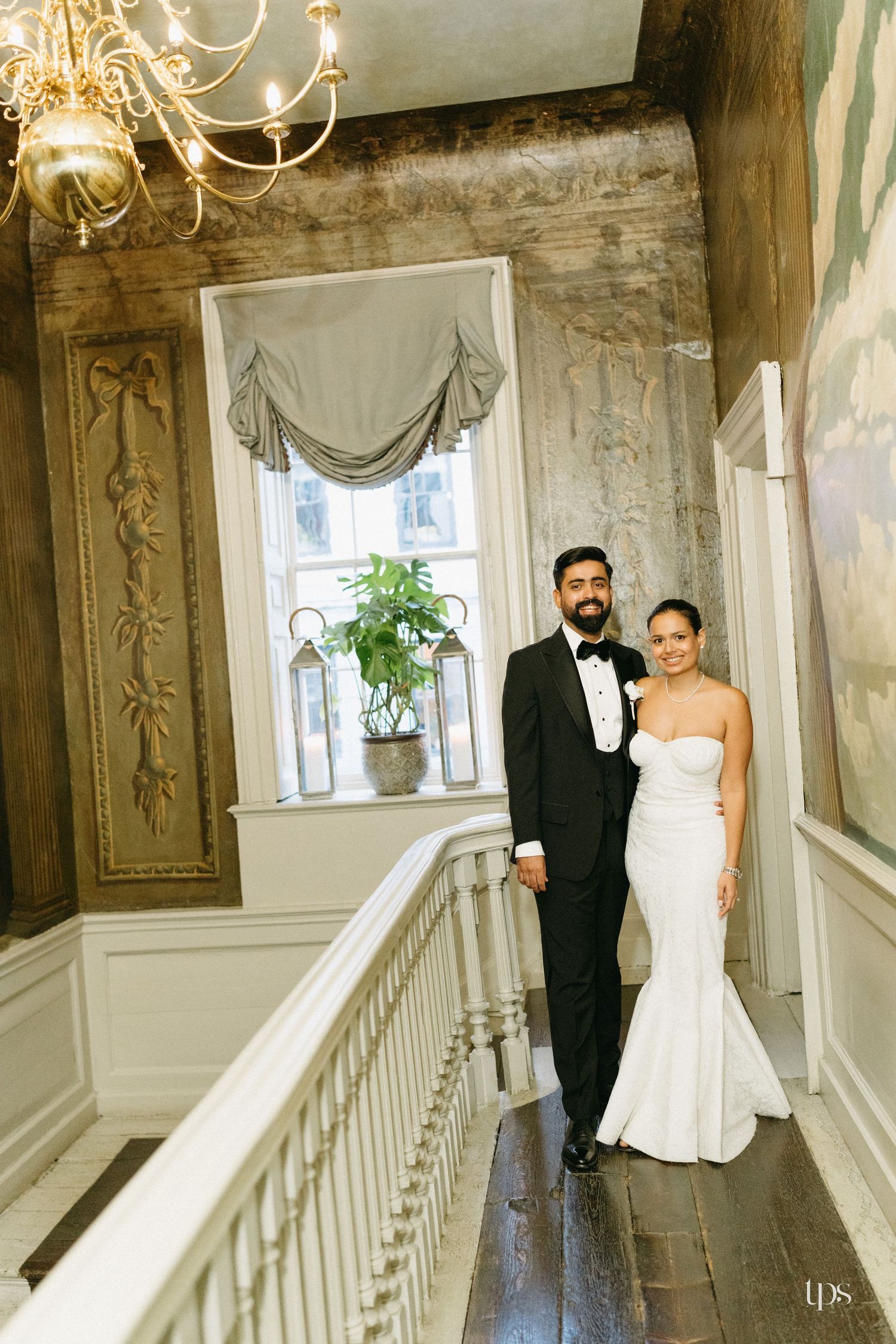 Bride and groom pose elegantly on grand staircase in ornate venue, she in fitted white gown, he in classic black tuxedo.