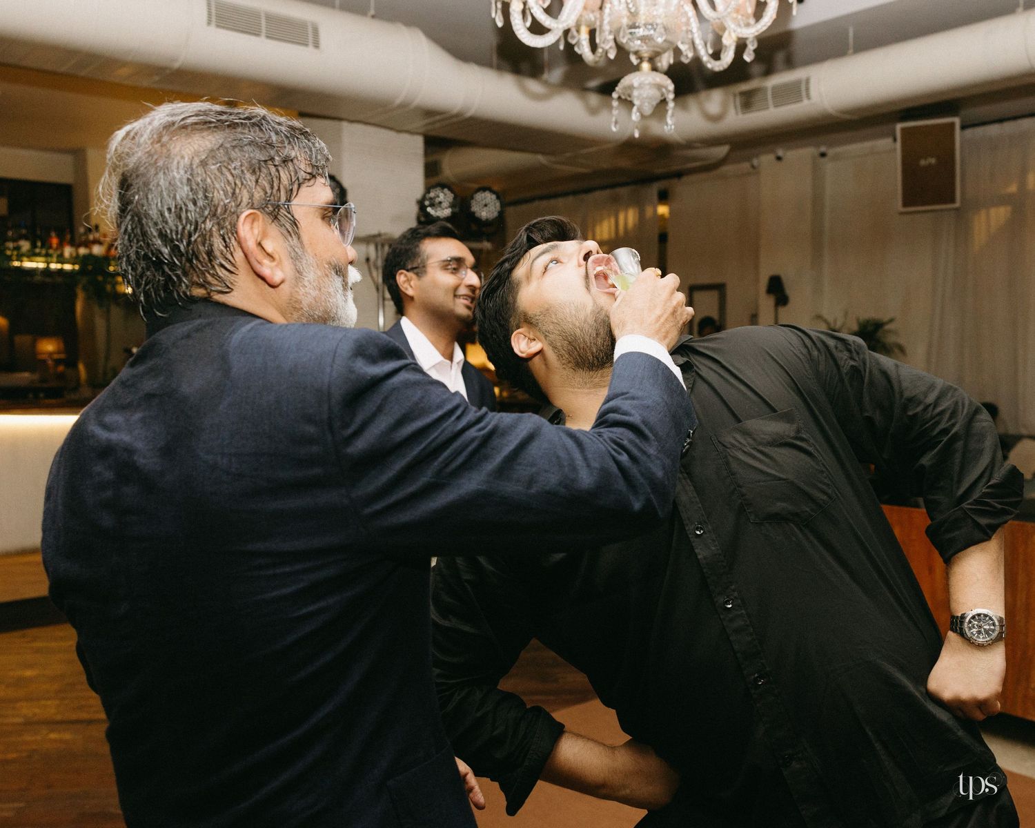Two men in dark shirts and a blazer laugh and drink at a lively indoor party while another guest looks on near a chandelier.