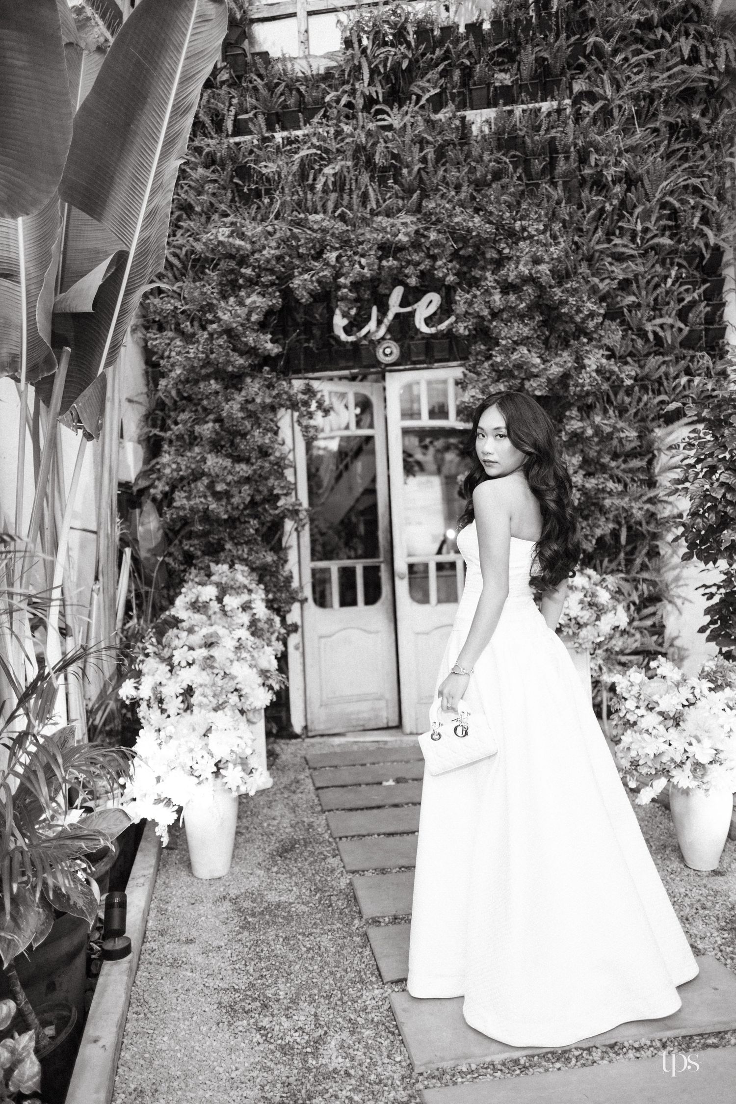 Black and white photo of a woman in a long strapless white gown holding a small handbag on a stone path before a vine-covered doorway.