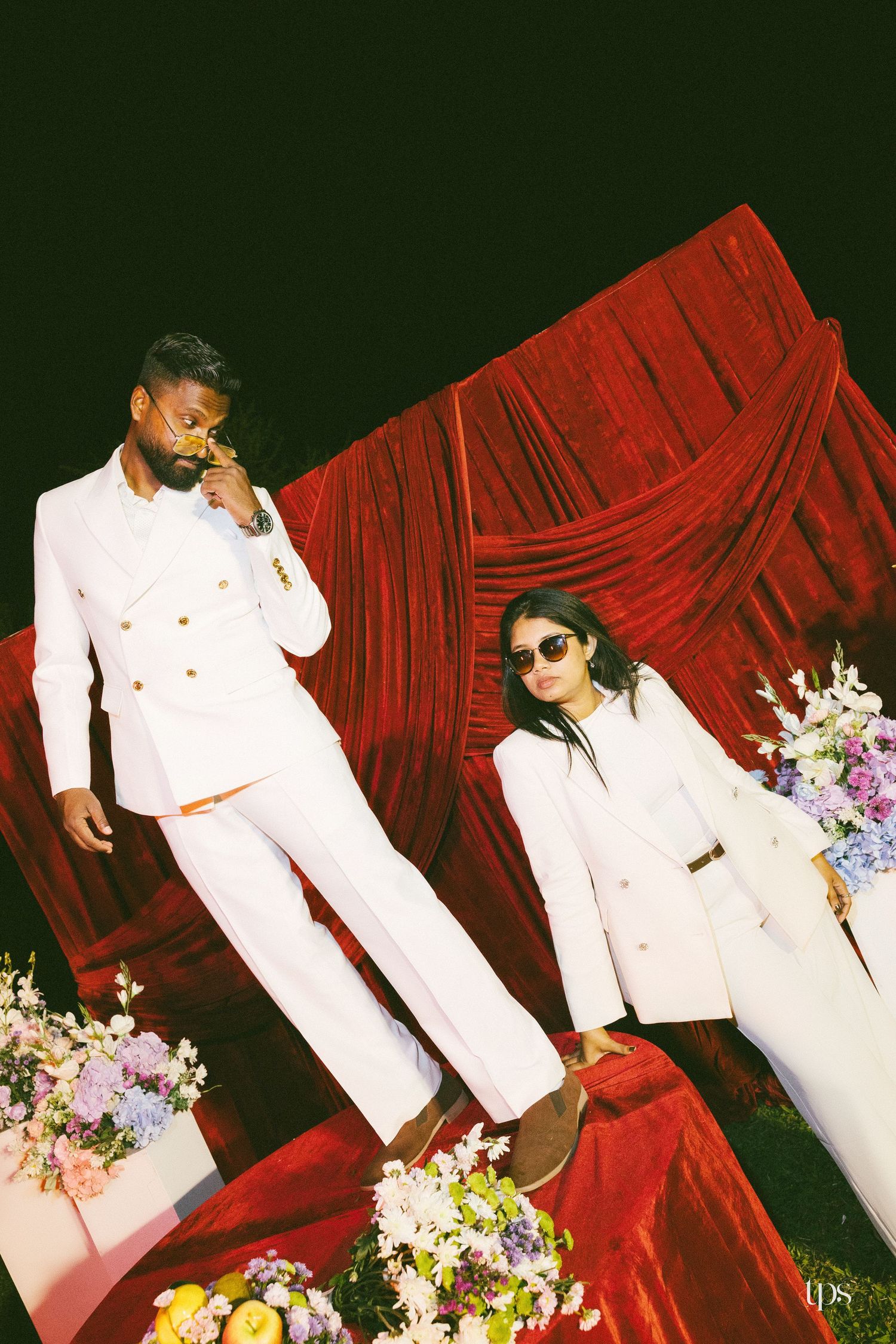 Two people in matching white double-breasted suits pose against a red velvet drape, surrounded by pastel floral arrangements and fruit.