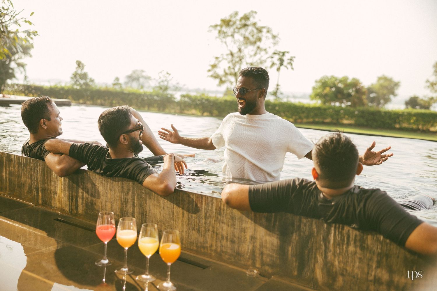 Four friends talk and laugh at the edge of an infinity pool with colorful cocktails lined up on the ledge, surrounded by lush green trees.