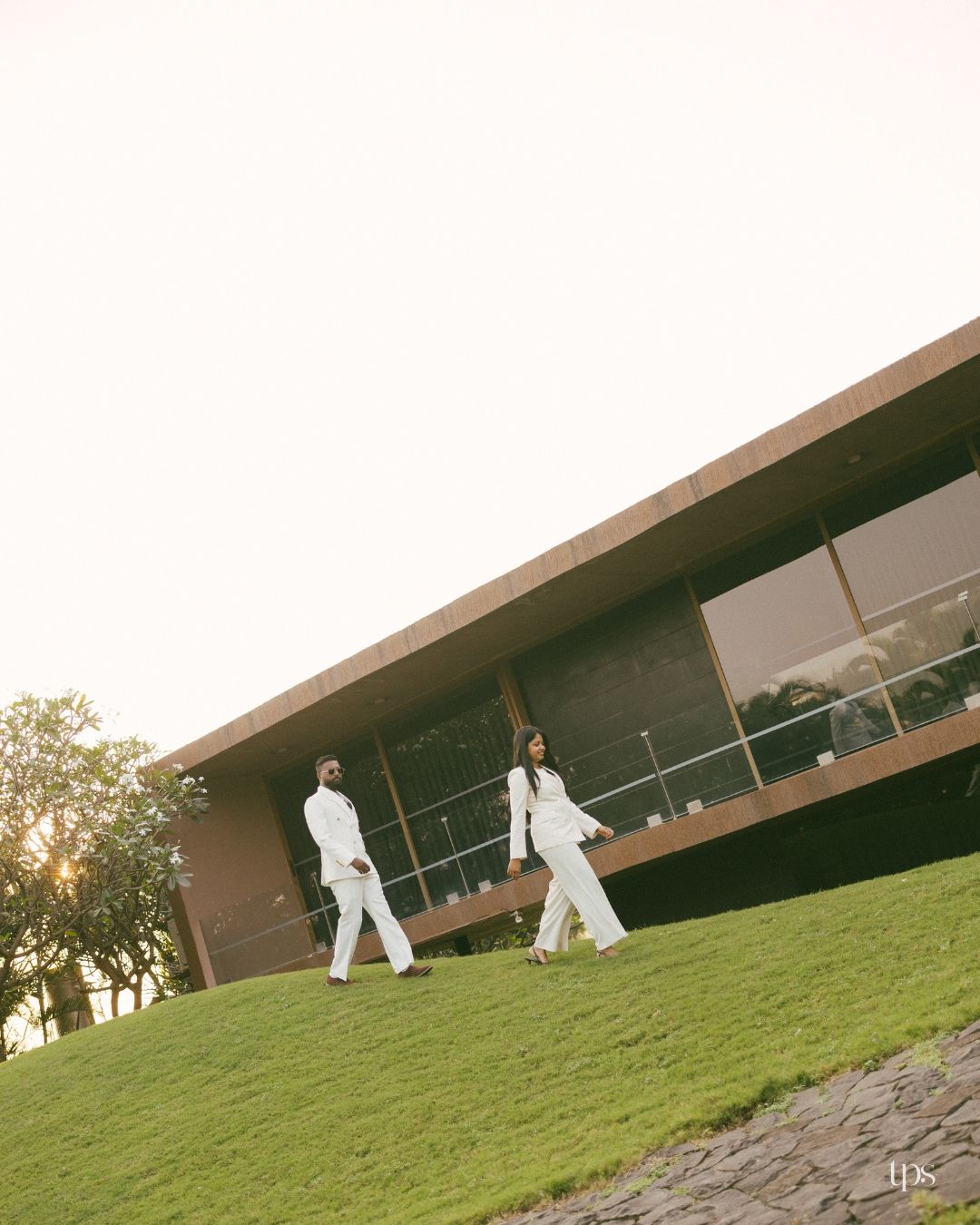 Couple in matching white suits walking along a green grassy hillside beside a modern brick and glass building at sunset.