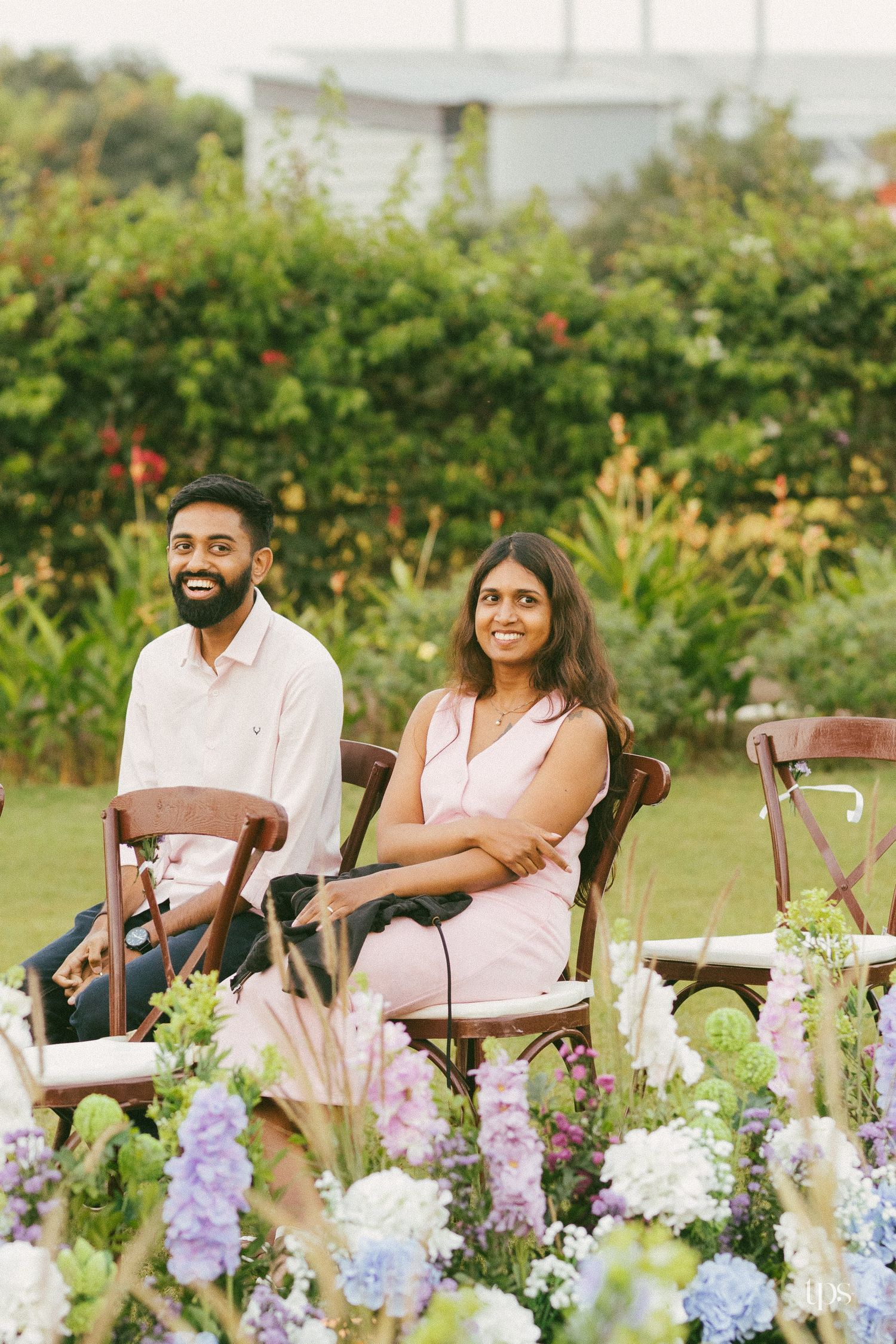 Smiling couple in pink and white outfits sit on wooden chairs in a lush garden with colorful hydrangea and floral arrangements.