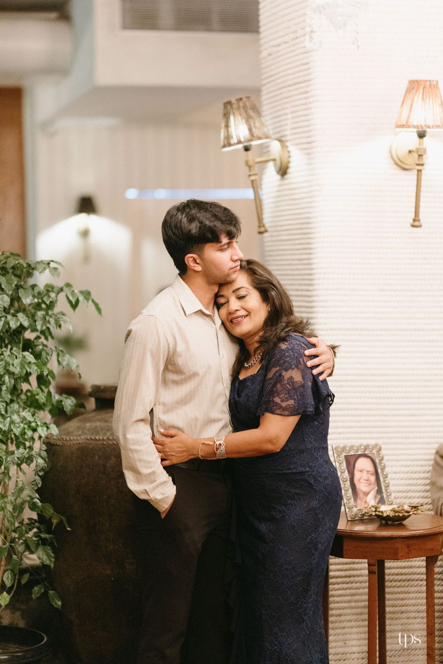 A young man in a cream shirt embraces a smiling woman in a navy lace dress beside a framed portrait and warm wall sconces.