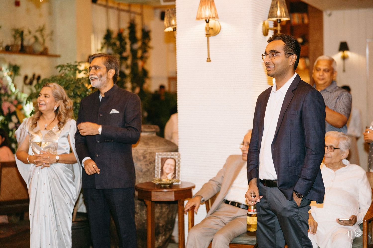 Guests in formal attire stand and sit in a warmly lit room with plants and wall sconces, listening to a speaker at an elegant gathering.