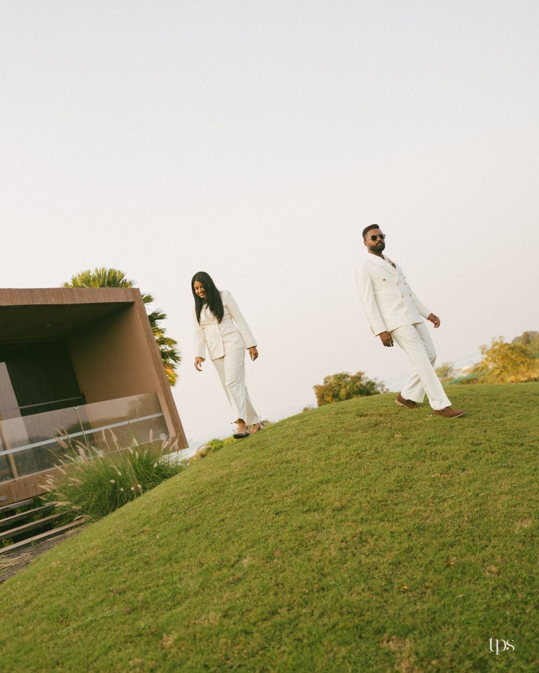 Two people in matching white suits walk down a green grassy hillside toward a modern building under a soft, hazy sky.