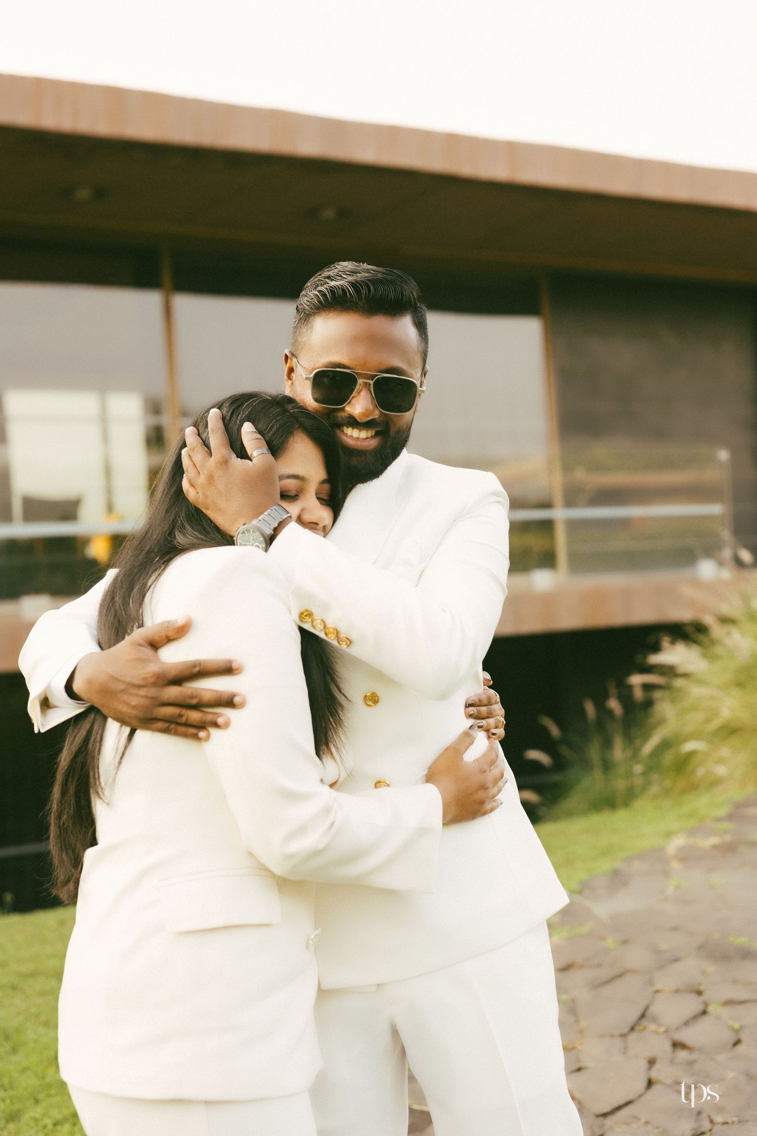 A smiling couple in matching white suits shares a warm embrace on a lawn outside a modern building during a pre-wedding photoshoot.
