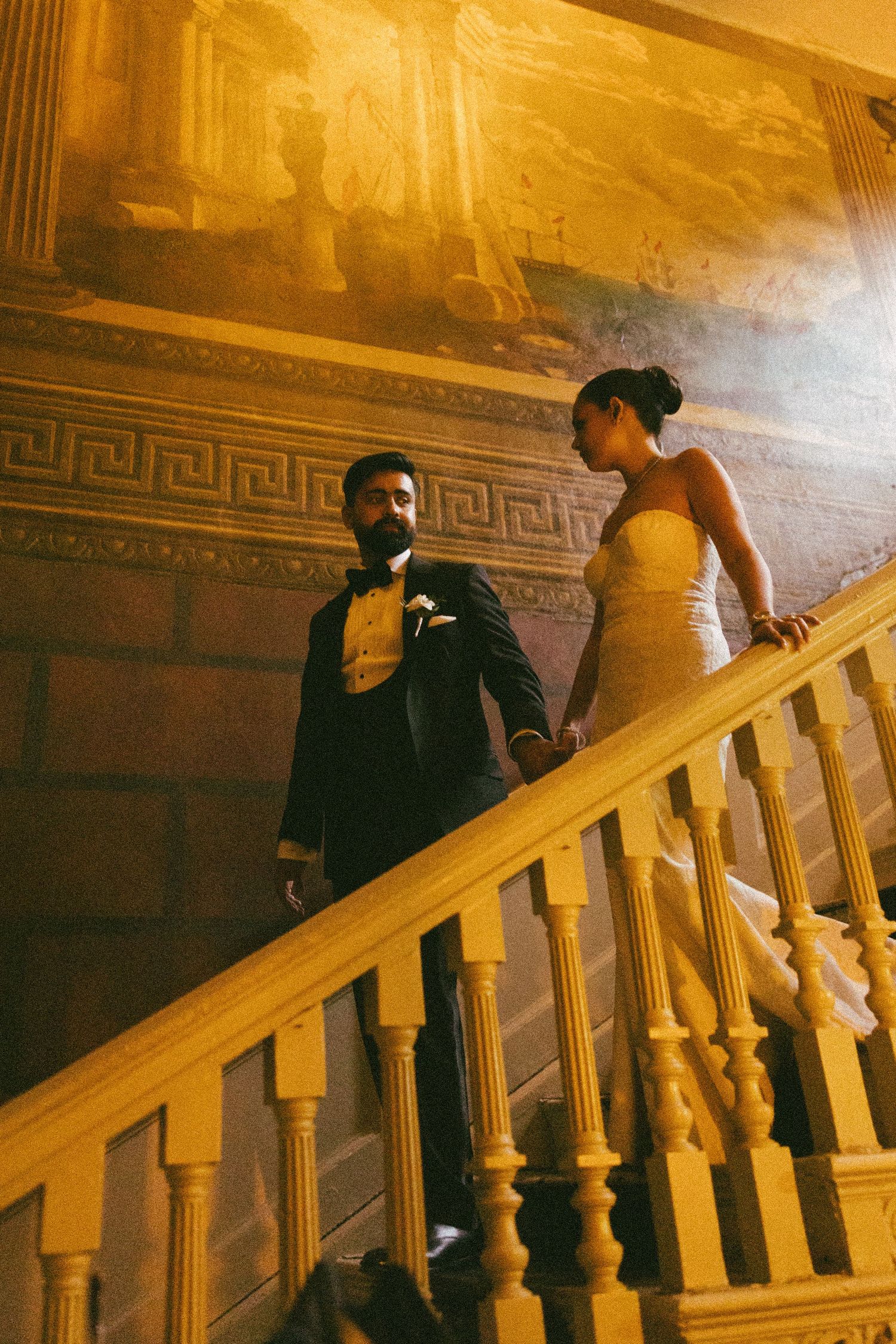 Elegant wedding couple descending ornate staircase, groom in tuxedo and bride in strapless gown, golden mural backdrop.