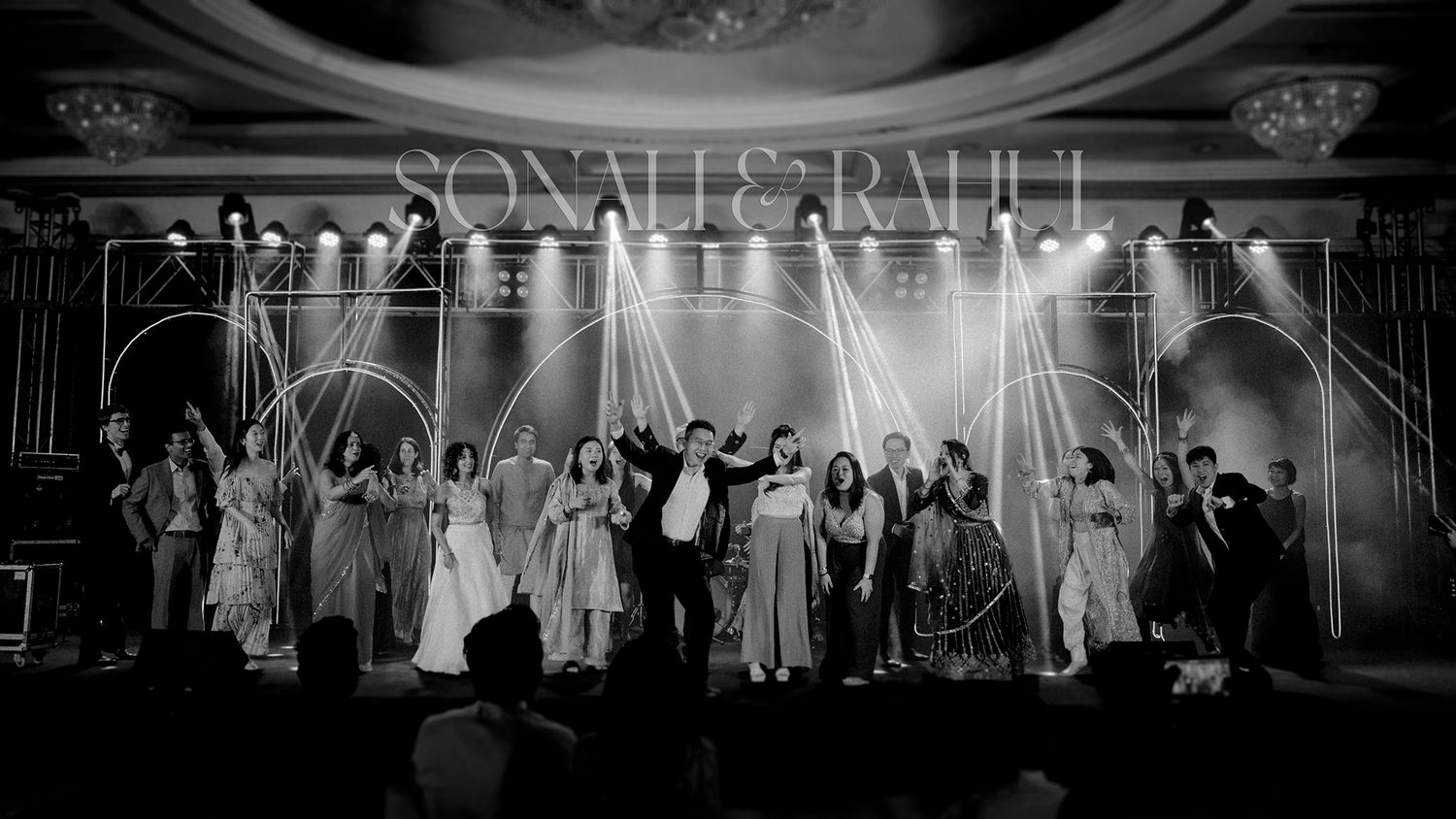 Black and white wedding reception photo of bride and groom on stage with wedding party under elegant arch decor.