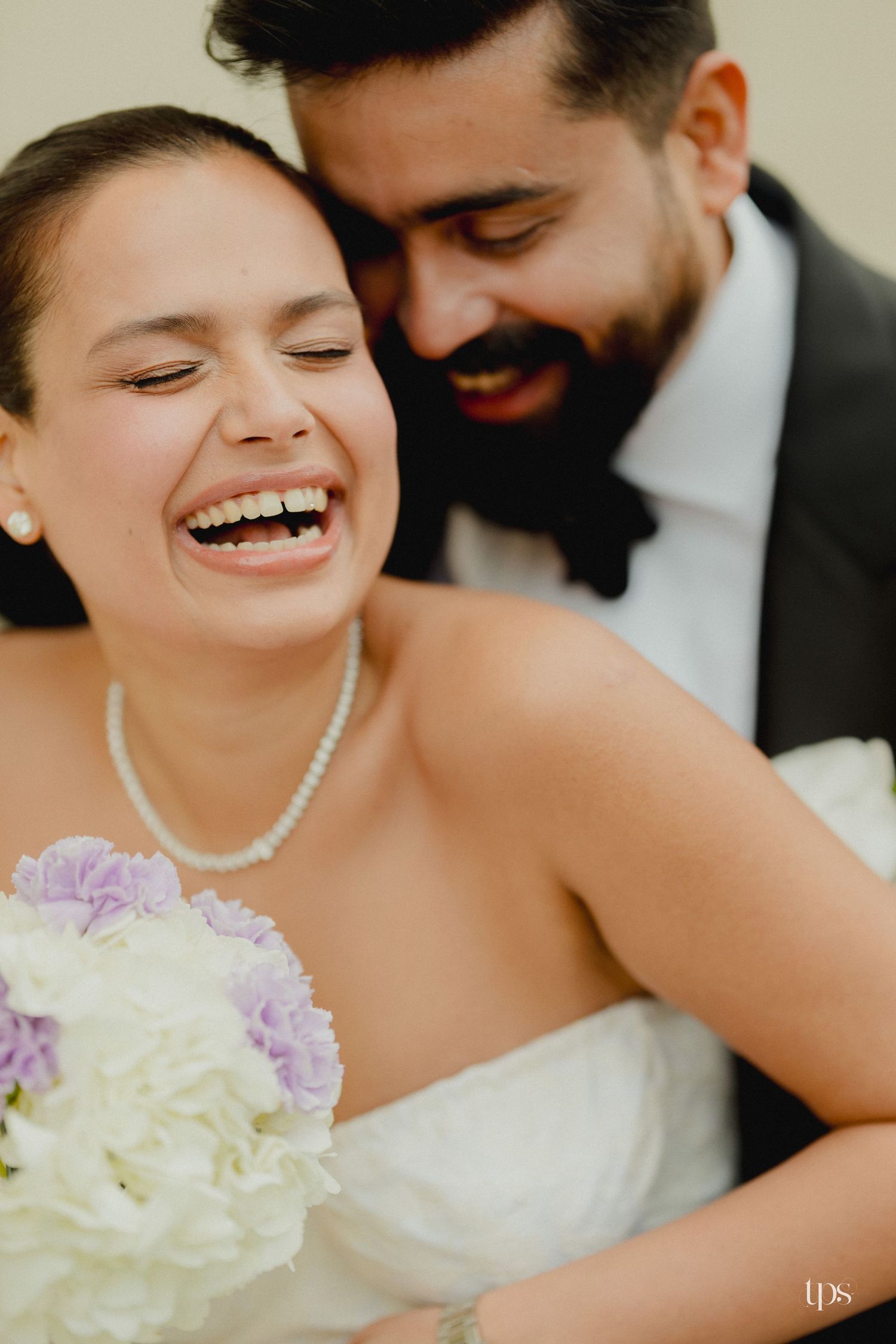 Joyful bride laughing with groom in tuxedo embracing her, holding white and lavender bouquet on wedding day.