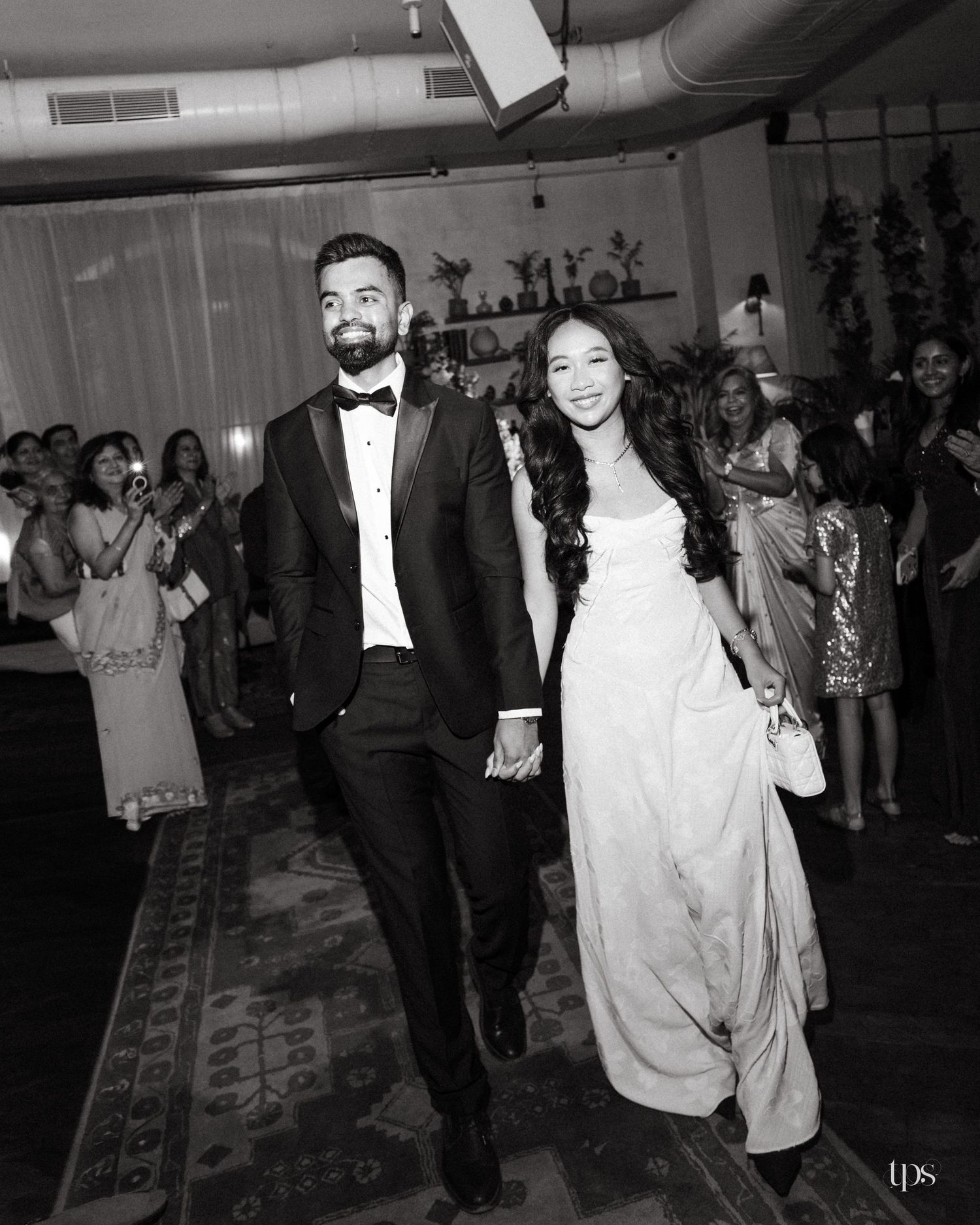 A smiling couple holds hands while walking through a reception as guests look on, the groom in a tuxedo and the bride in a flowing gown.