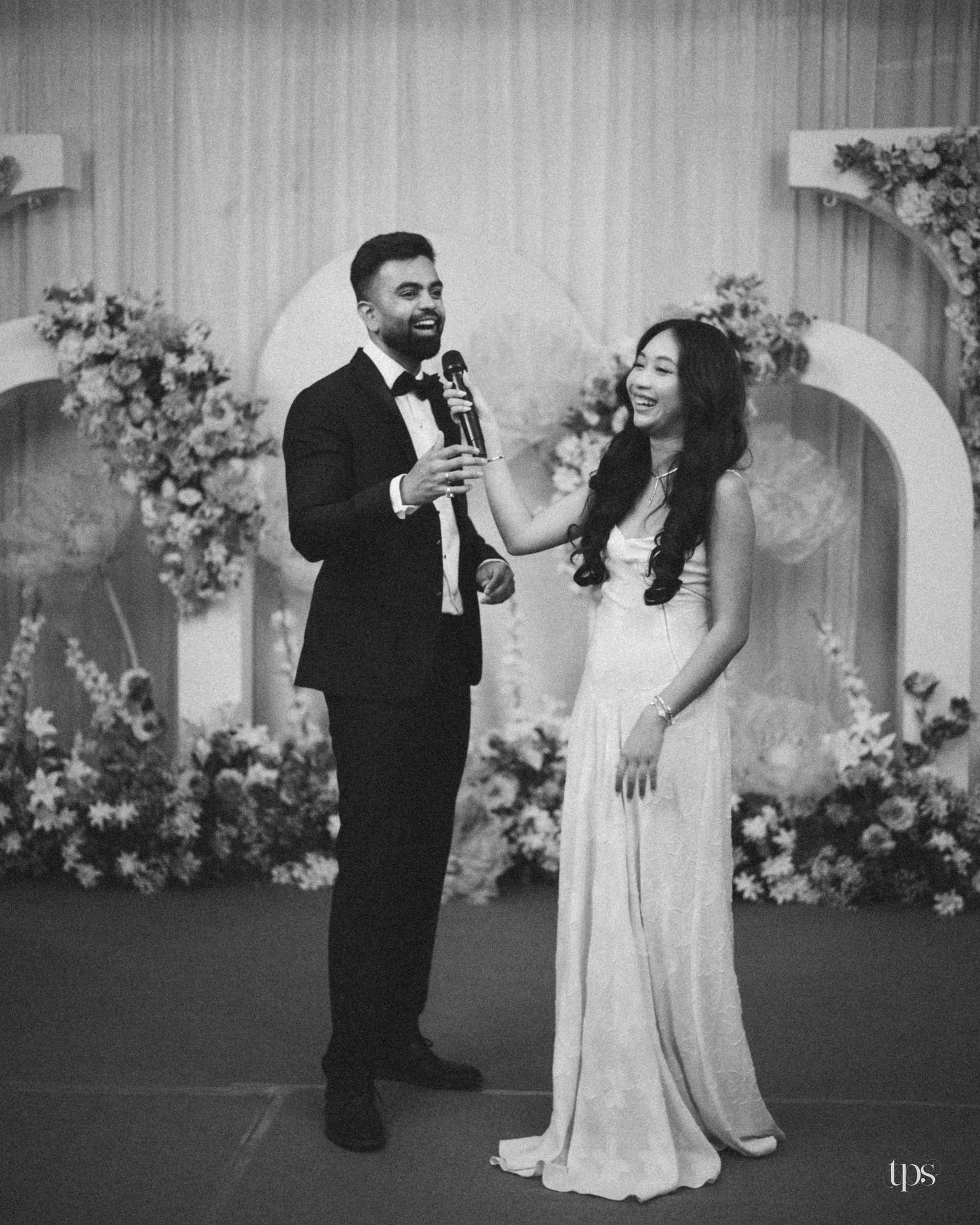 Black and white photo of a groom in a tuxedo and a bride in a flowing gown sharing a microphone at a wedding reception with floral arches.