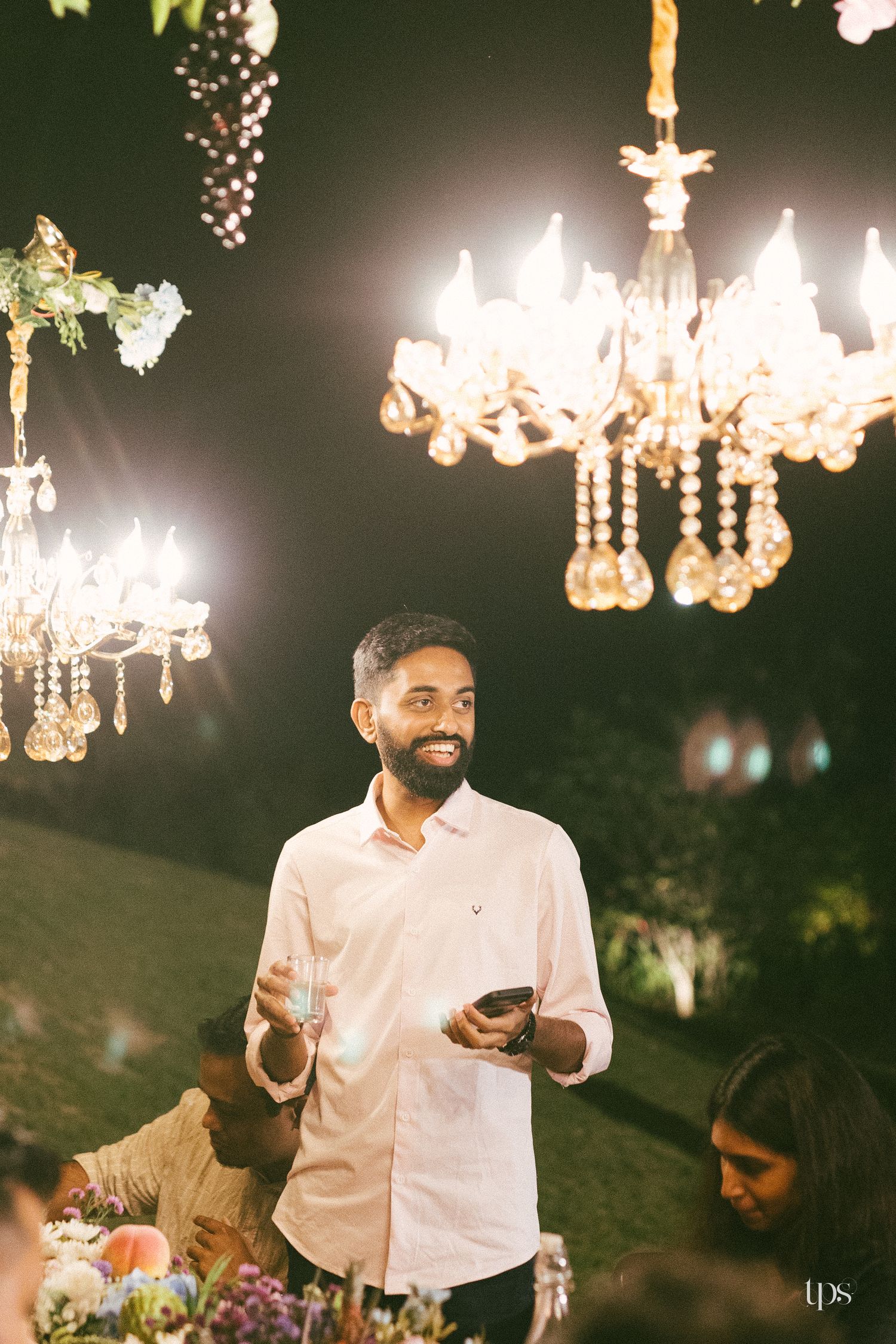 Smiling bearded man in a pink shirt holds a glass and phone at an outdoor evening event lit by ornate crystal chandeliers.
