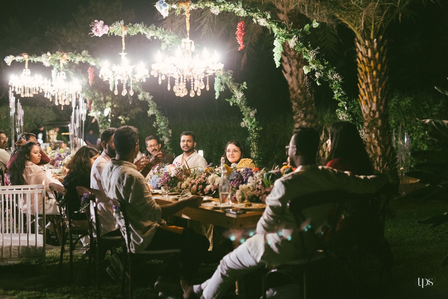 Guests gather around a long candlelit table beneath sparkling chandeliers and greenery at an elegant outdoor evening garden dinner party.