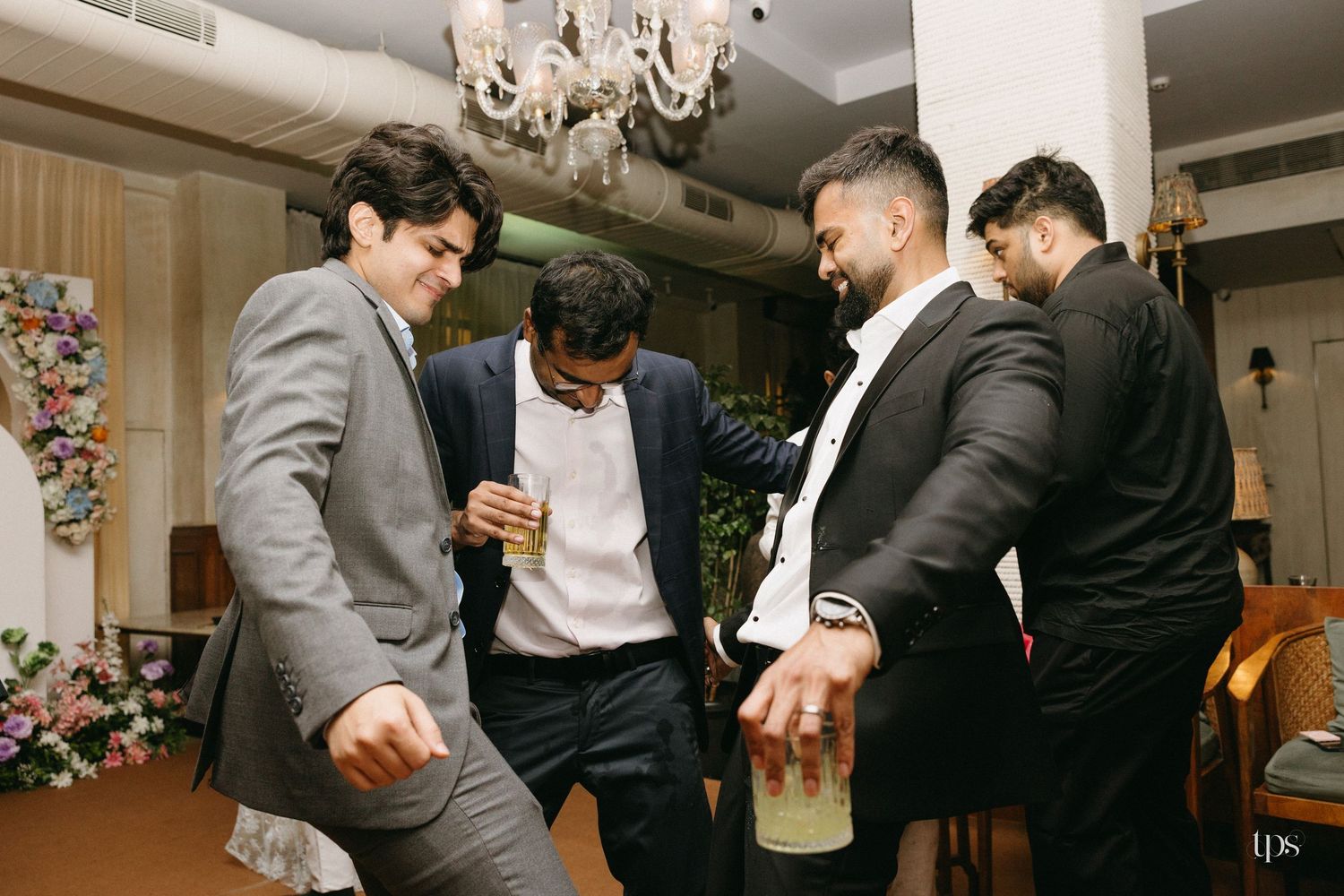 Four men in suits laugh and dance while holding drinks at an indoor celebration with a chandelier and floral decor.