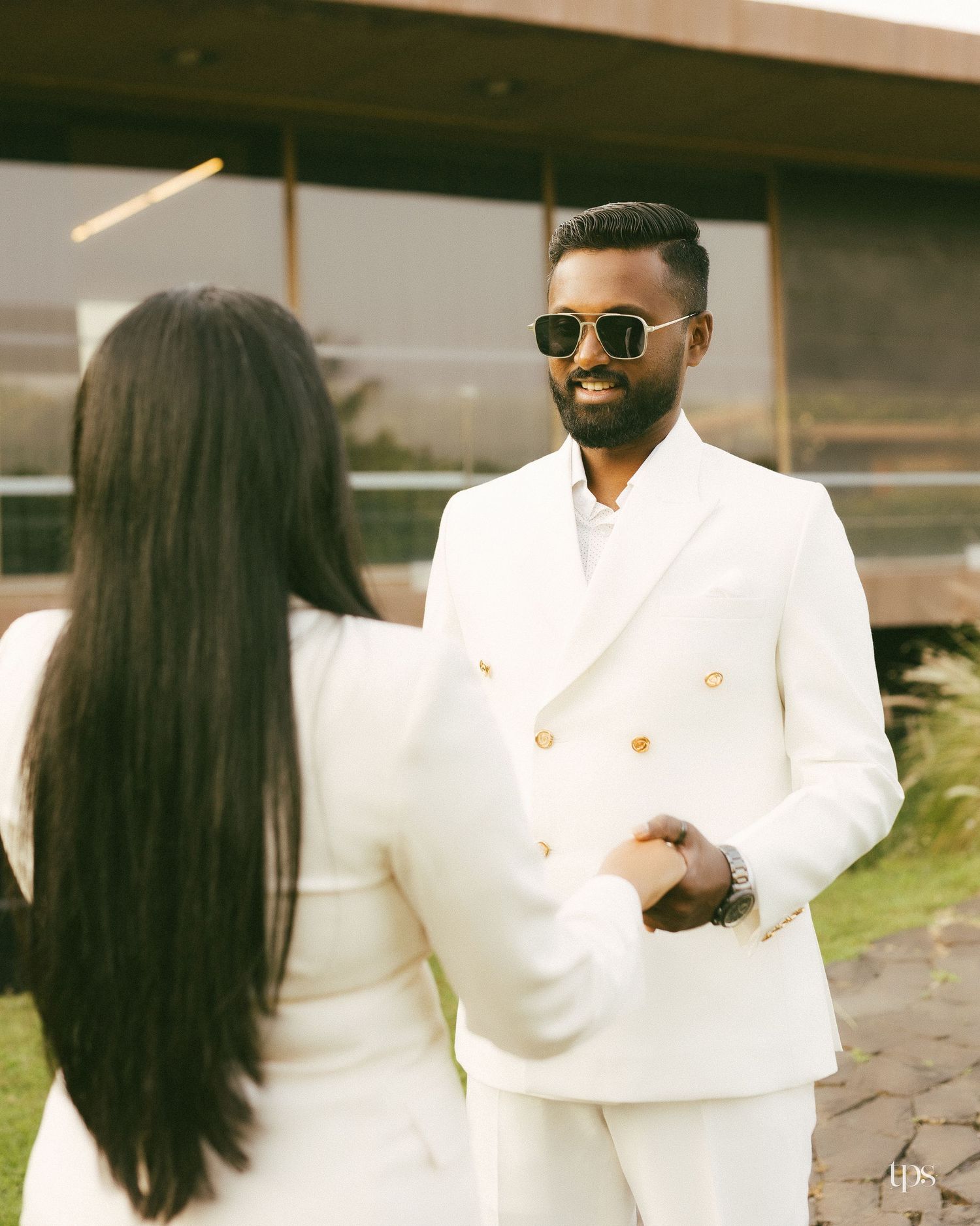 A bearded man in sunglasses and a white double-breasted suit holds hands with a long-haired woman in white outdoors.