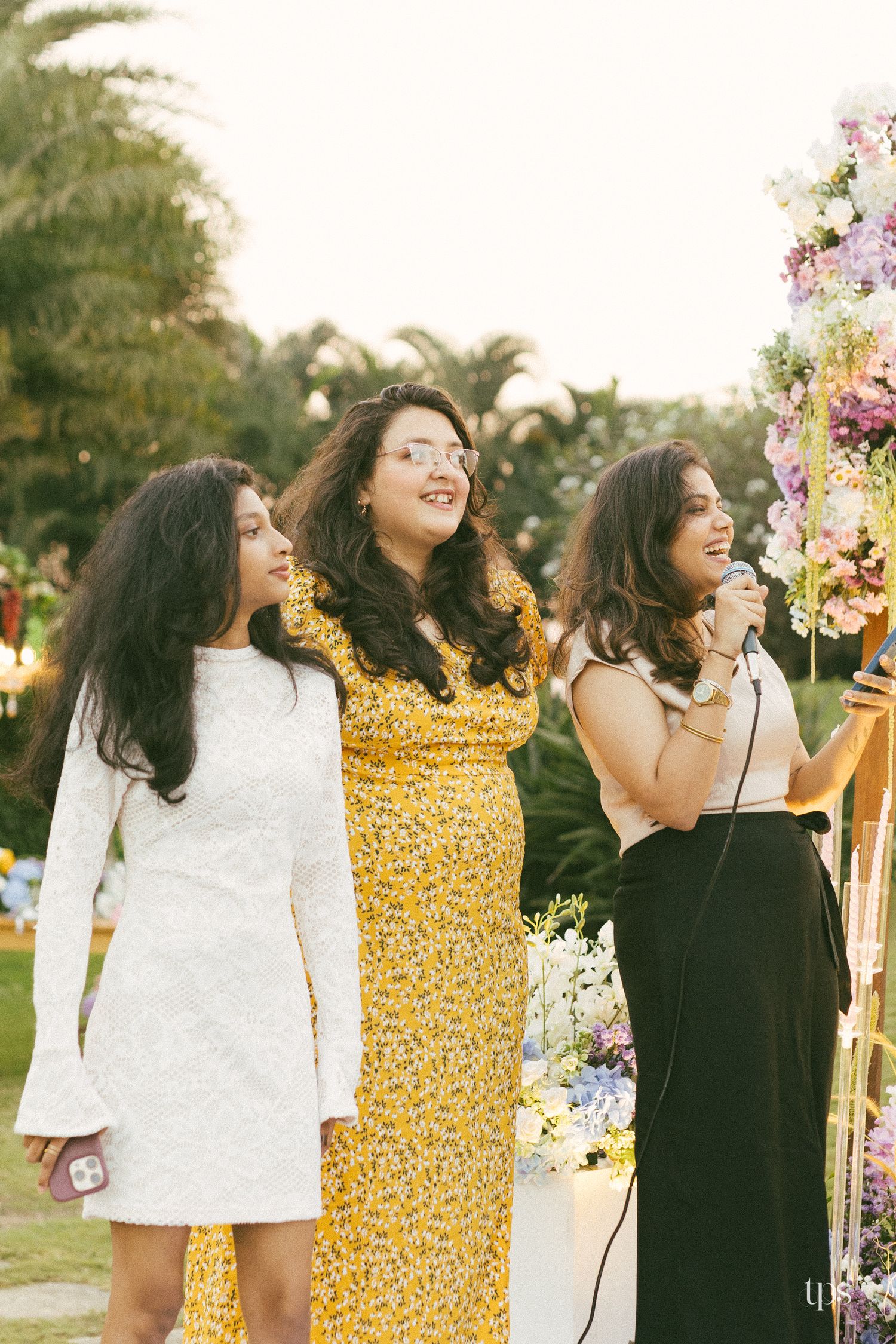 Three women in dresses stand together at an outdoor garden event, one singing into a microphone beside colorful flower arrangements.