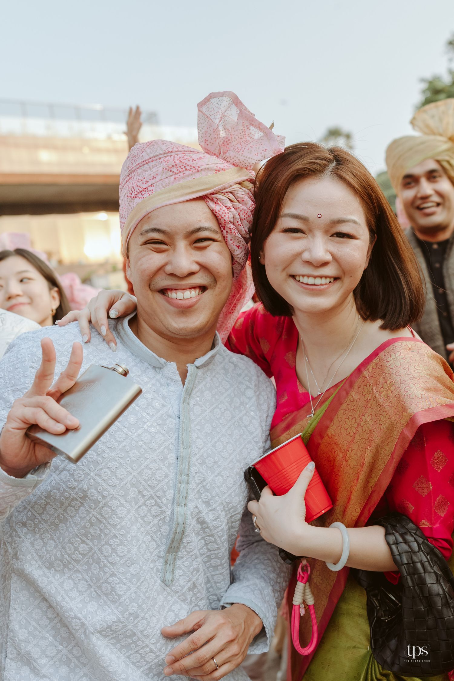 A smiling couple poses at an Indian wedding, man wearing pink turban and white kurta, woman in colorful silk saree.