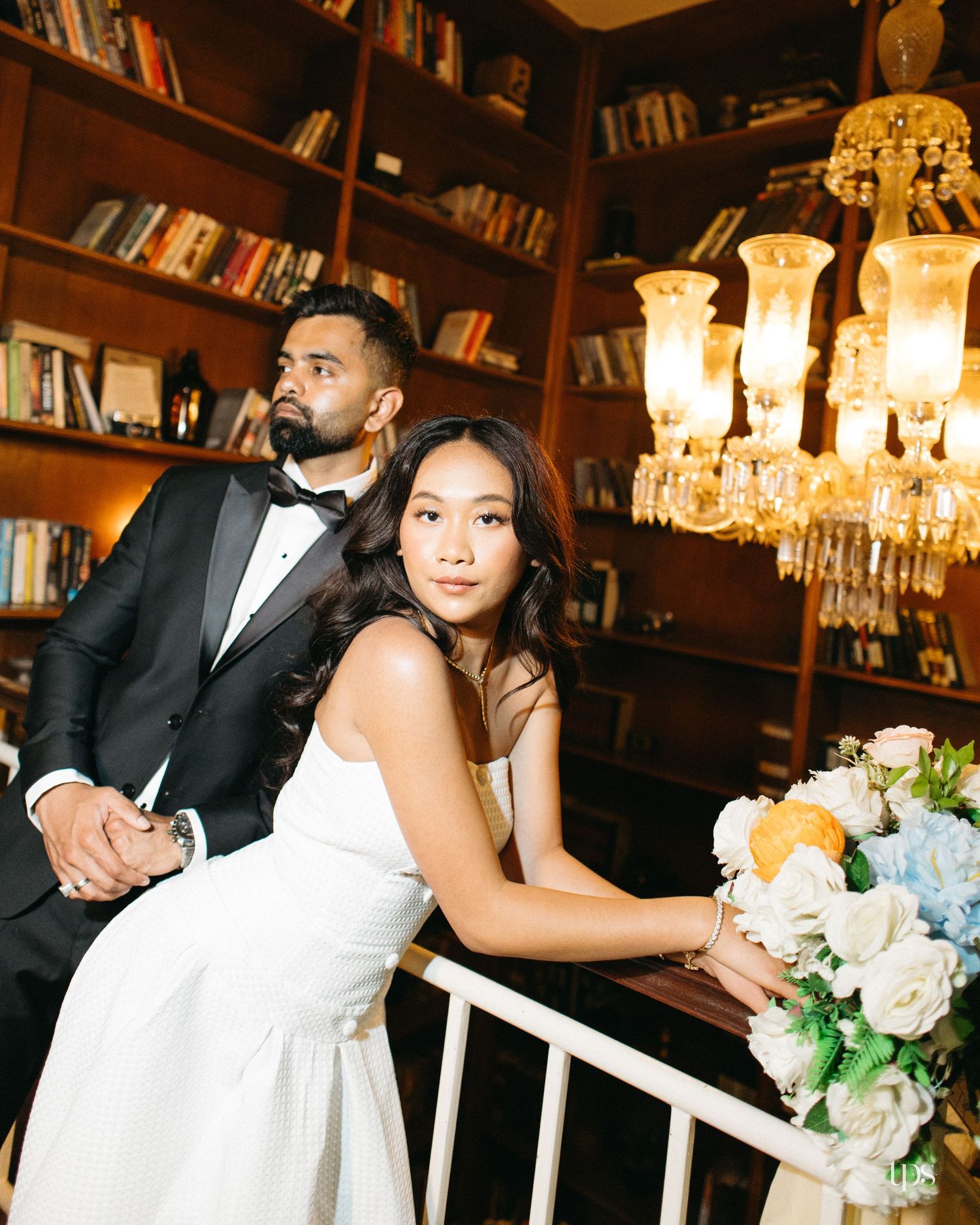 Bride in a white dress and groom in a black tuxedo pose by a staircase railing in a library with a crystal chandelier and a floral bouquet.