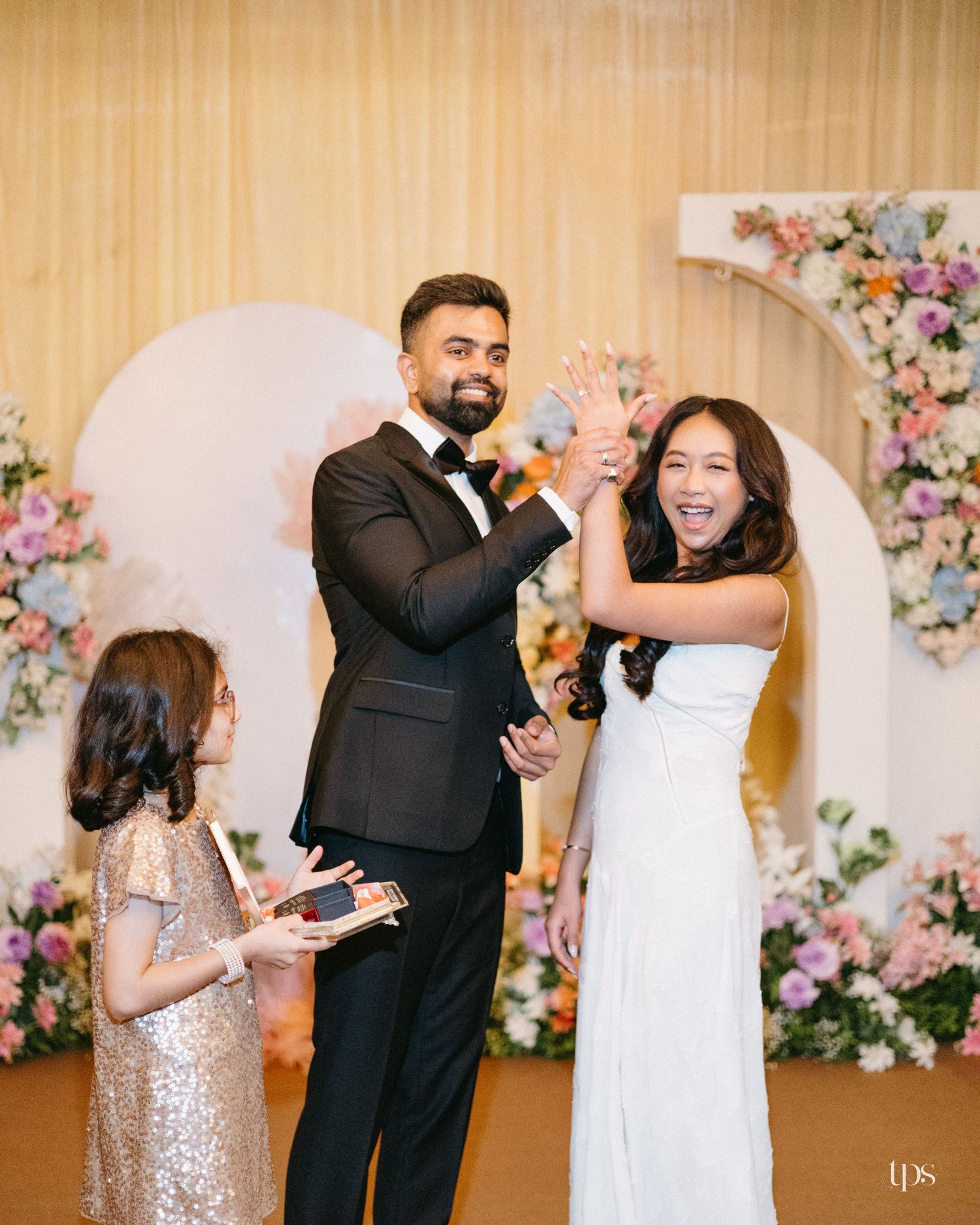 A smiling couple in a black tuxedo and white gown shows their hands beside a girl in a sparkly dress at a floral wedding backdrop.